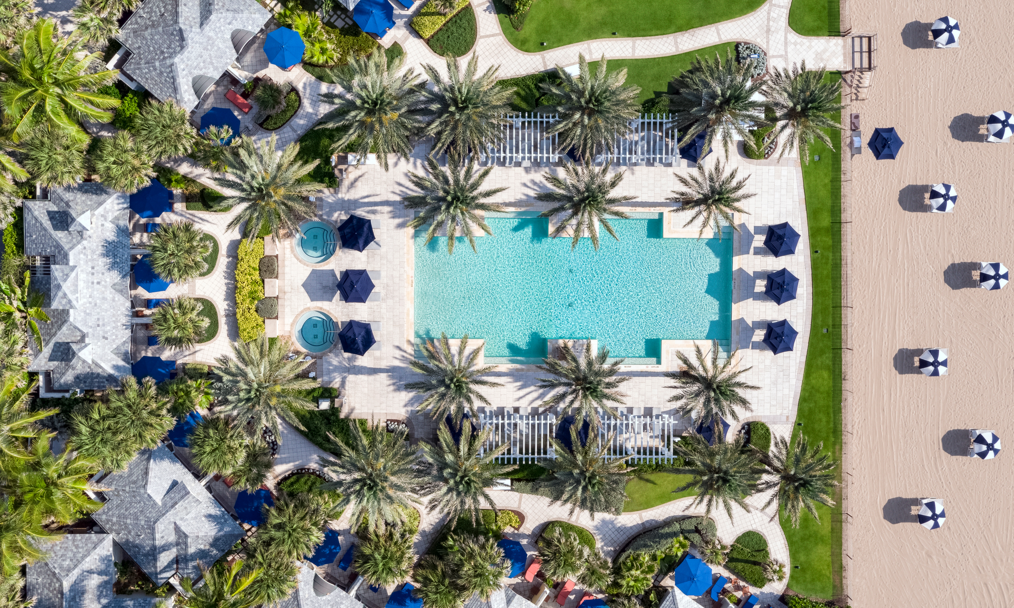 Aerial view of resort pool surrounded by palm trees, blue umbrellas, and lounge chairs on deck.