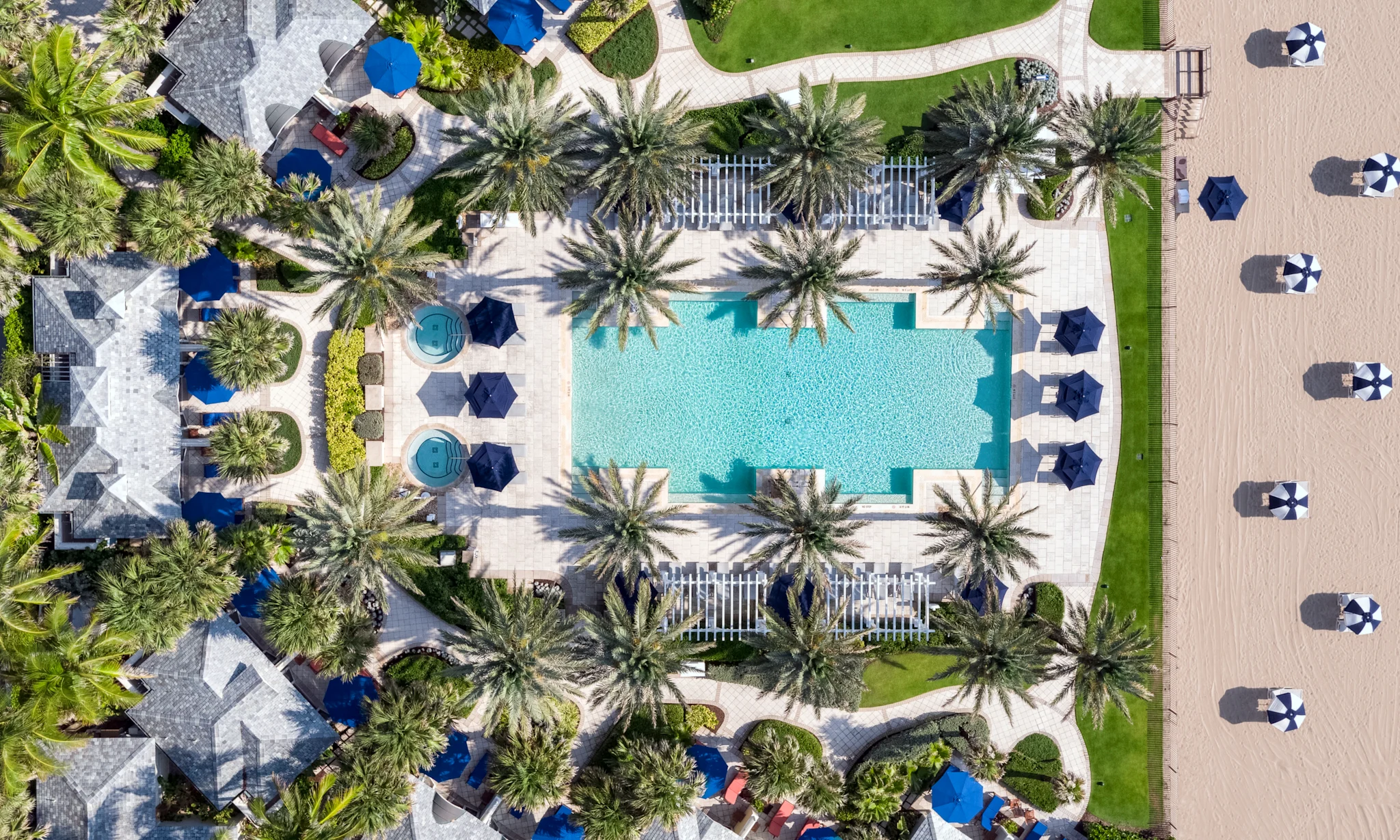 Aerial view of resort pool surrounded by palm trees, blue umbrellas, and lounge chairs on deck.