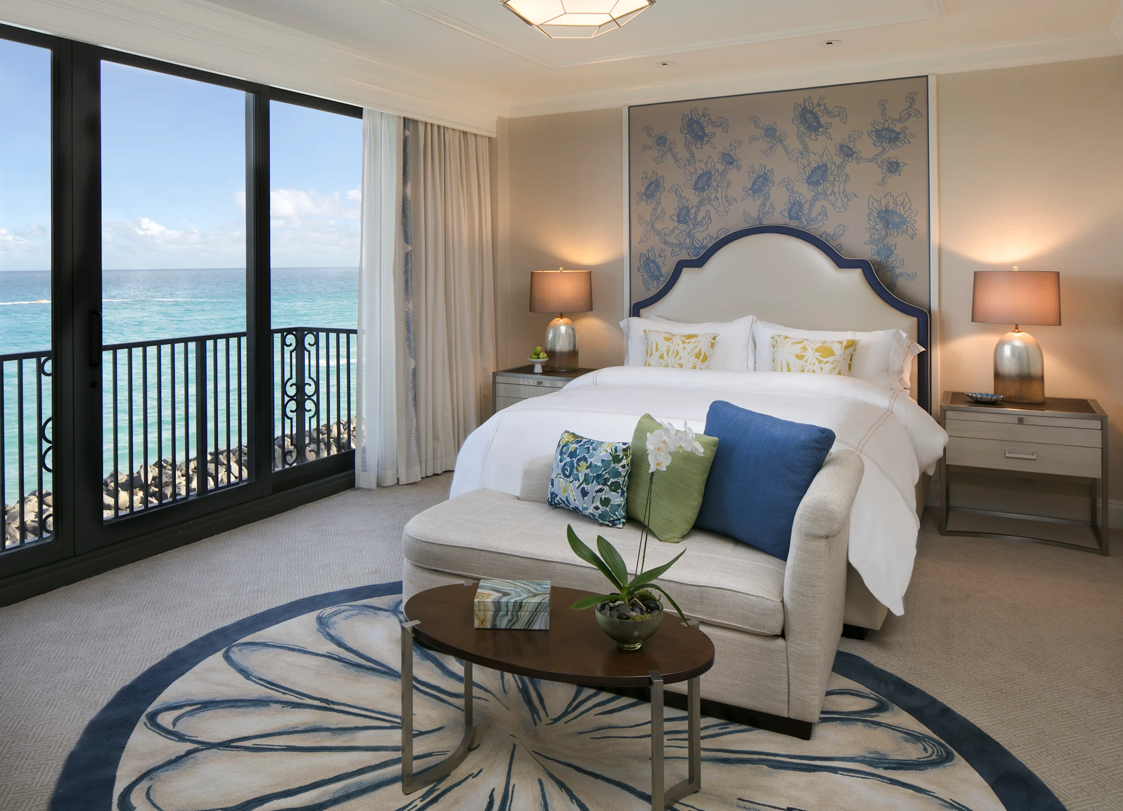 Elegant hotel bedroom with ocean view, white bed, blue accents, and curved sofa with decorative pillows.