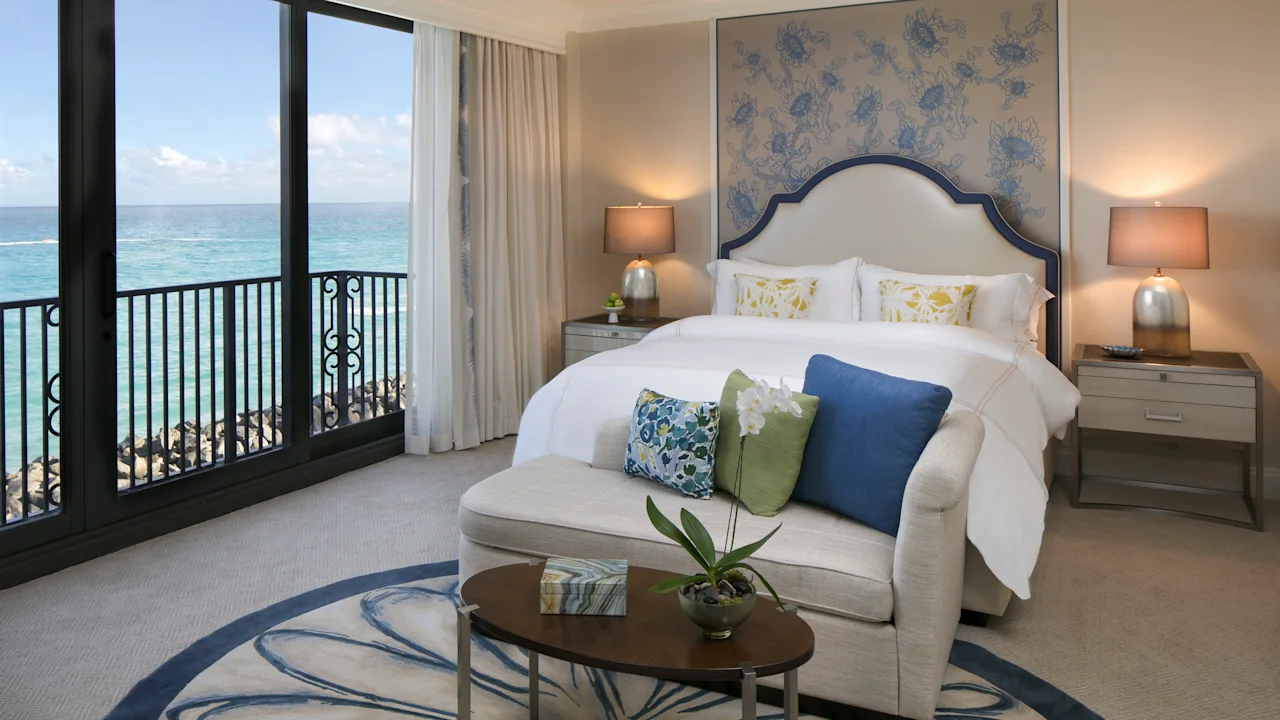 Elegant hotel bedroom with ocean view, white bed, blue accents, and curved sofa with decorative pillows.