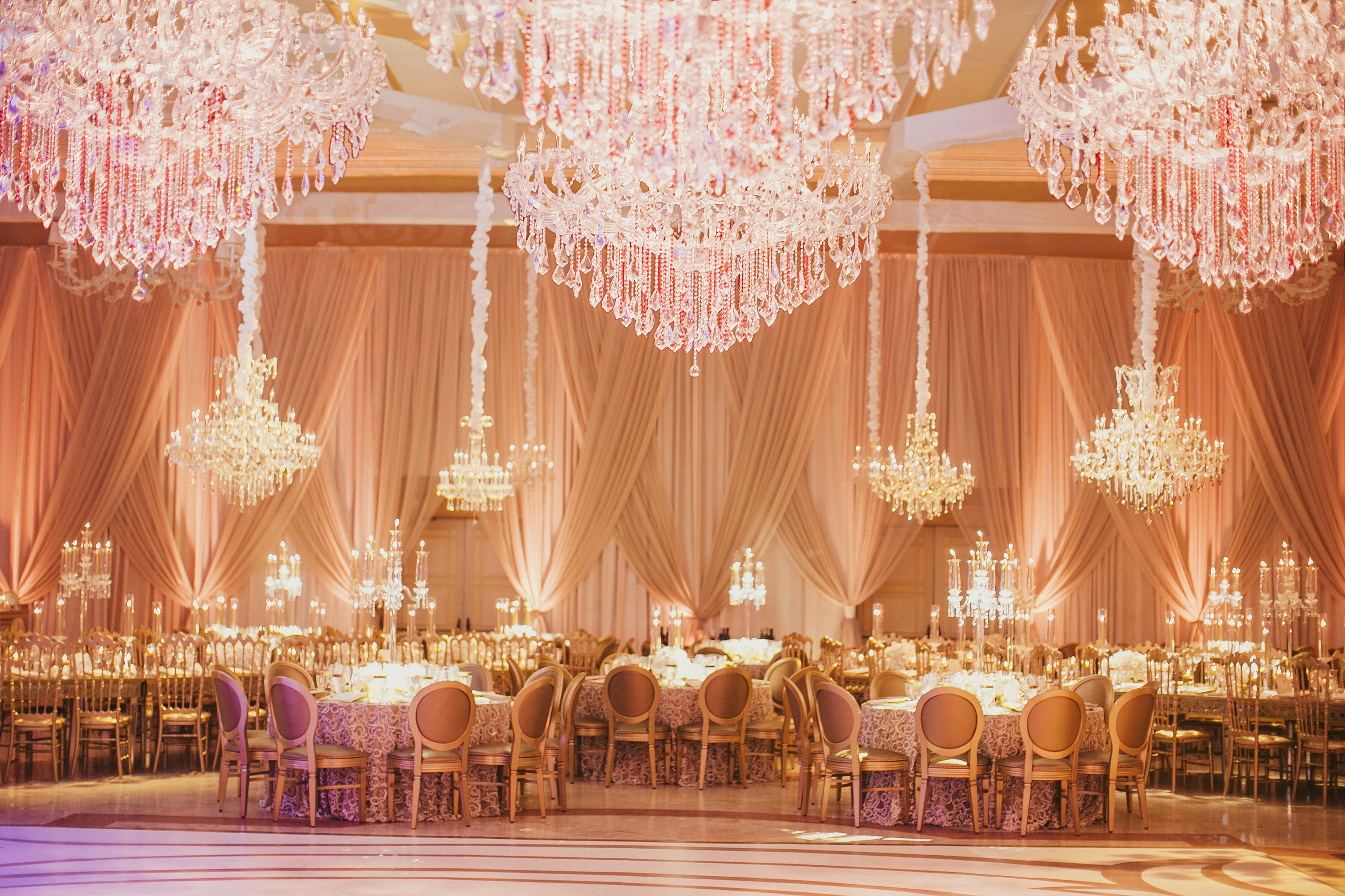 Elegant ballroom with crystal chandeliers, peach draping, and formal dining tables set for a wedding.