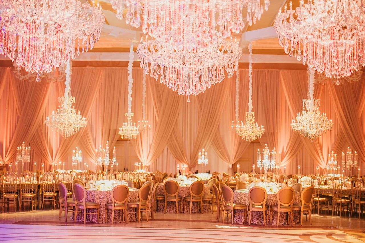 Elegant ballroom with crystal chandeliers, peach draping, and formal dining tables set for a wedding.
