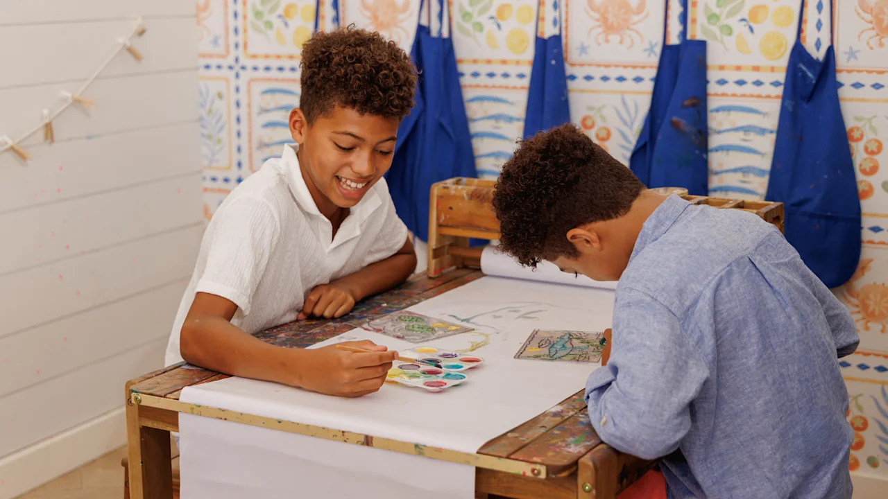 Two children painting together at a table with colorful tiles and blue aprons in the background.