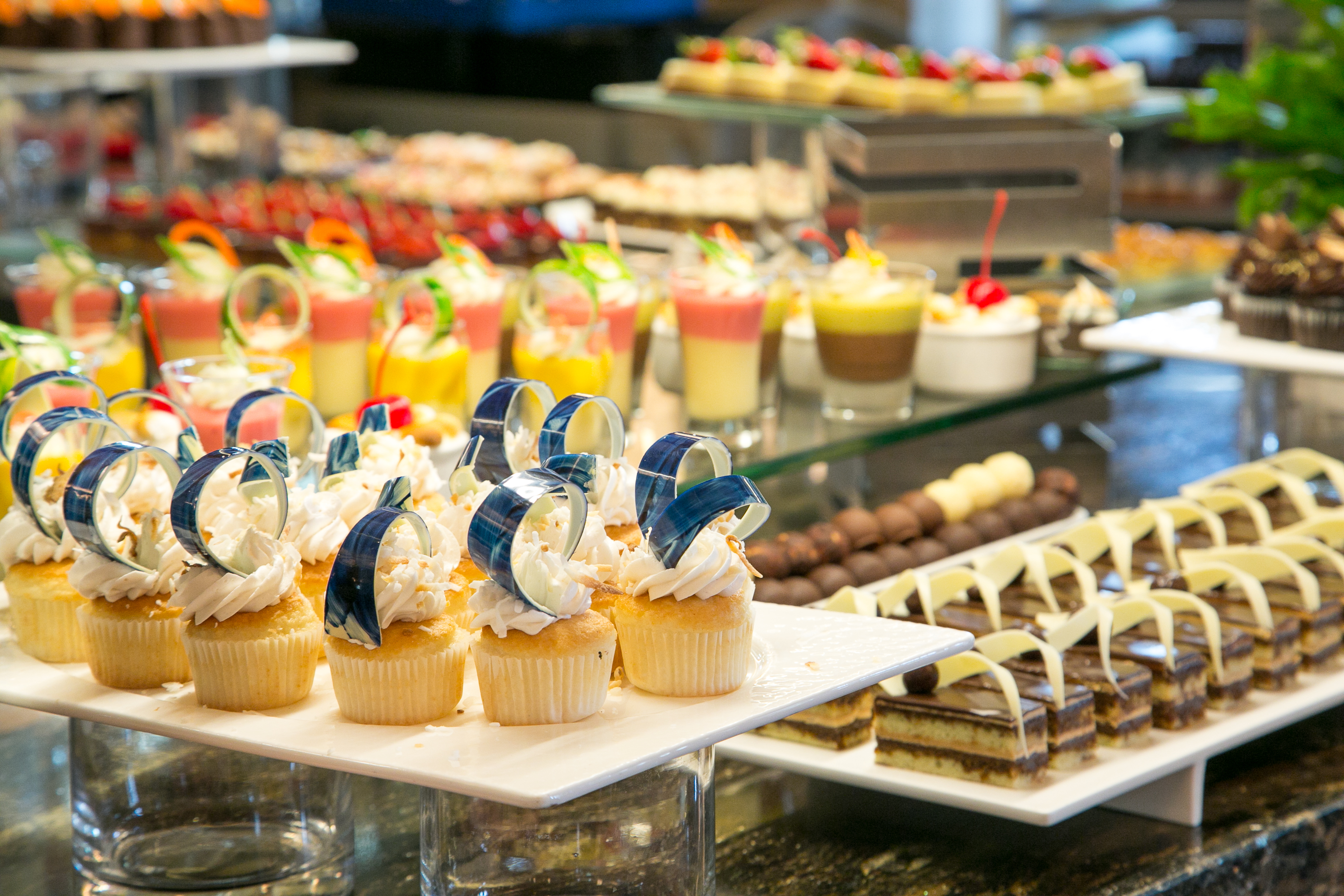 Dessert buffet display with decorated cupcakes, layered desserts, and colorful pastries on white trays.