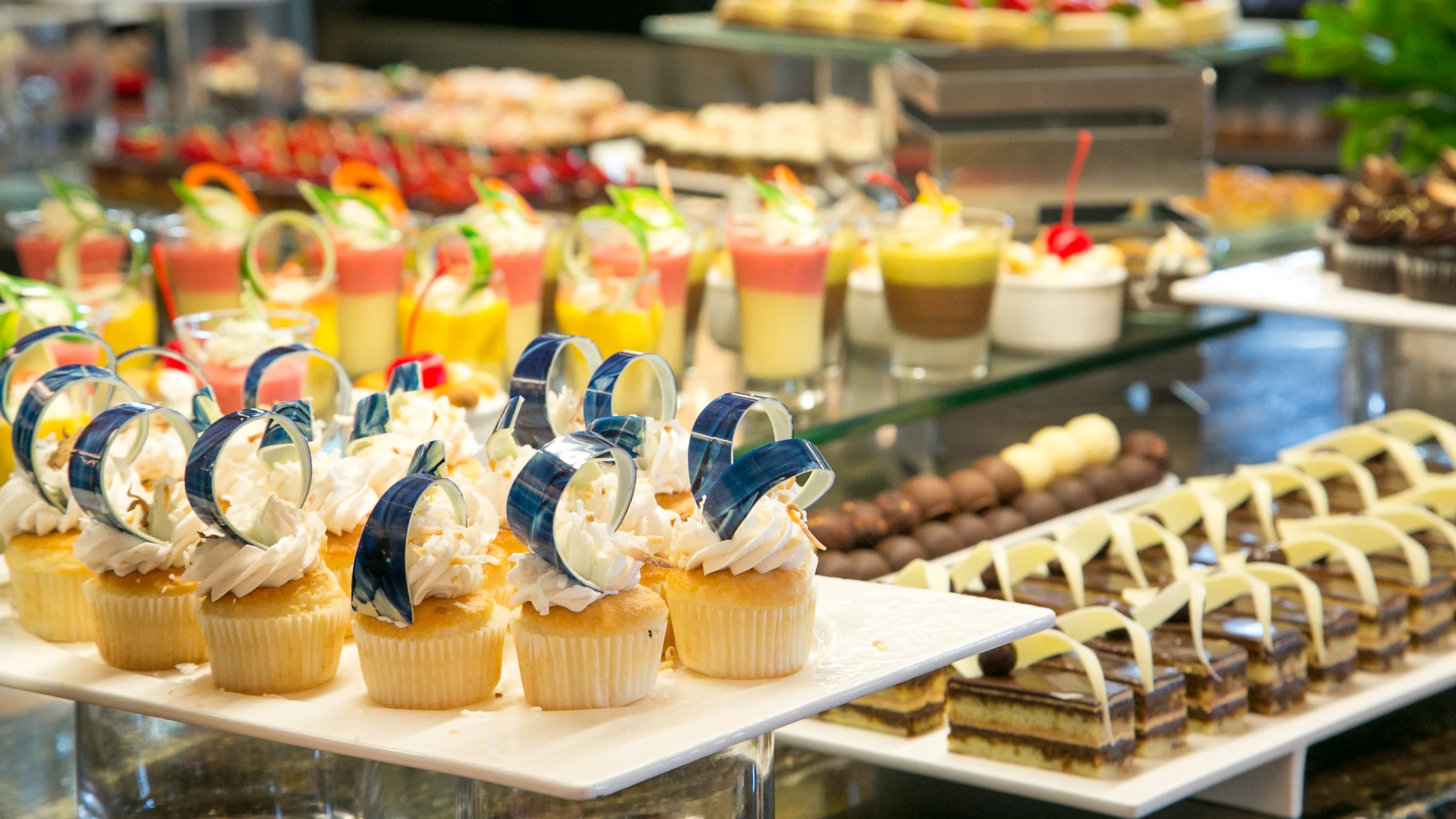 Dessert buffet display with decorated cupcakes, layered desserts, and colorful pastries on white trays.
