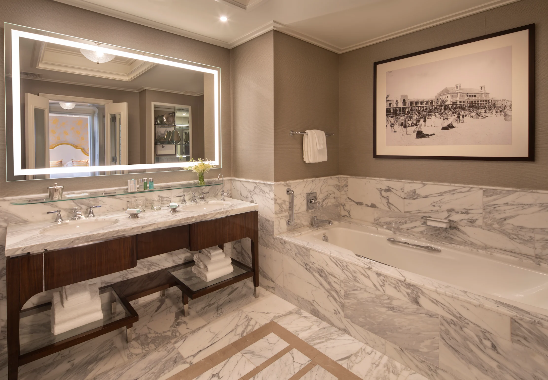 Luxury hotel bathroom with marble tub, double vanity, backlit mirror, and vintage beach photograph.