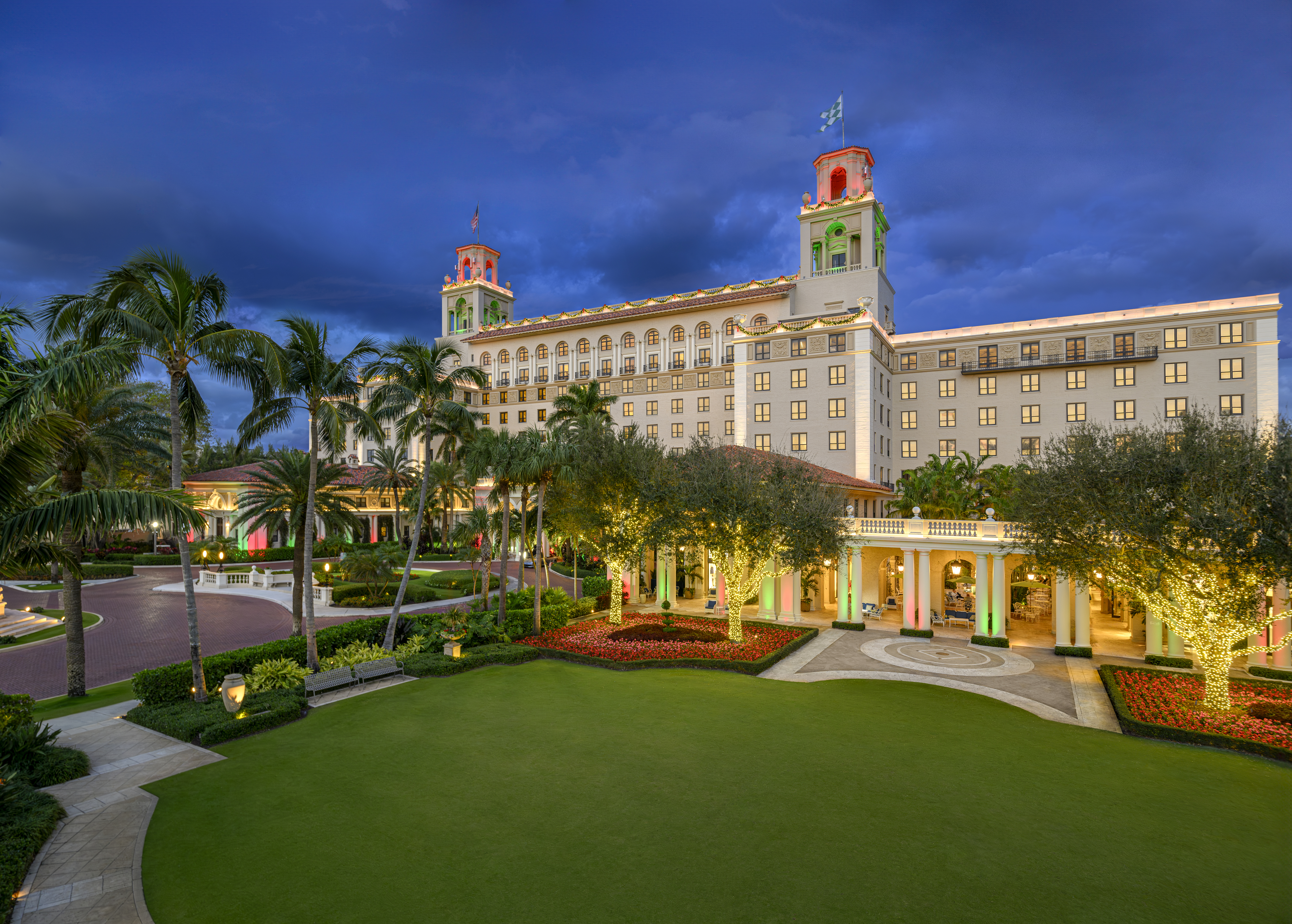Illuminated luxury hotel with twin towers at dusk, surrounded by palm trees and manicured lawns.