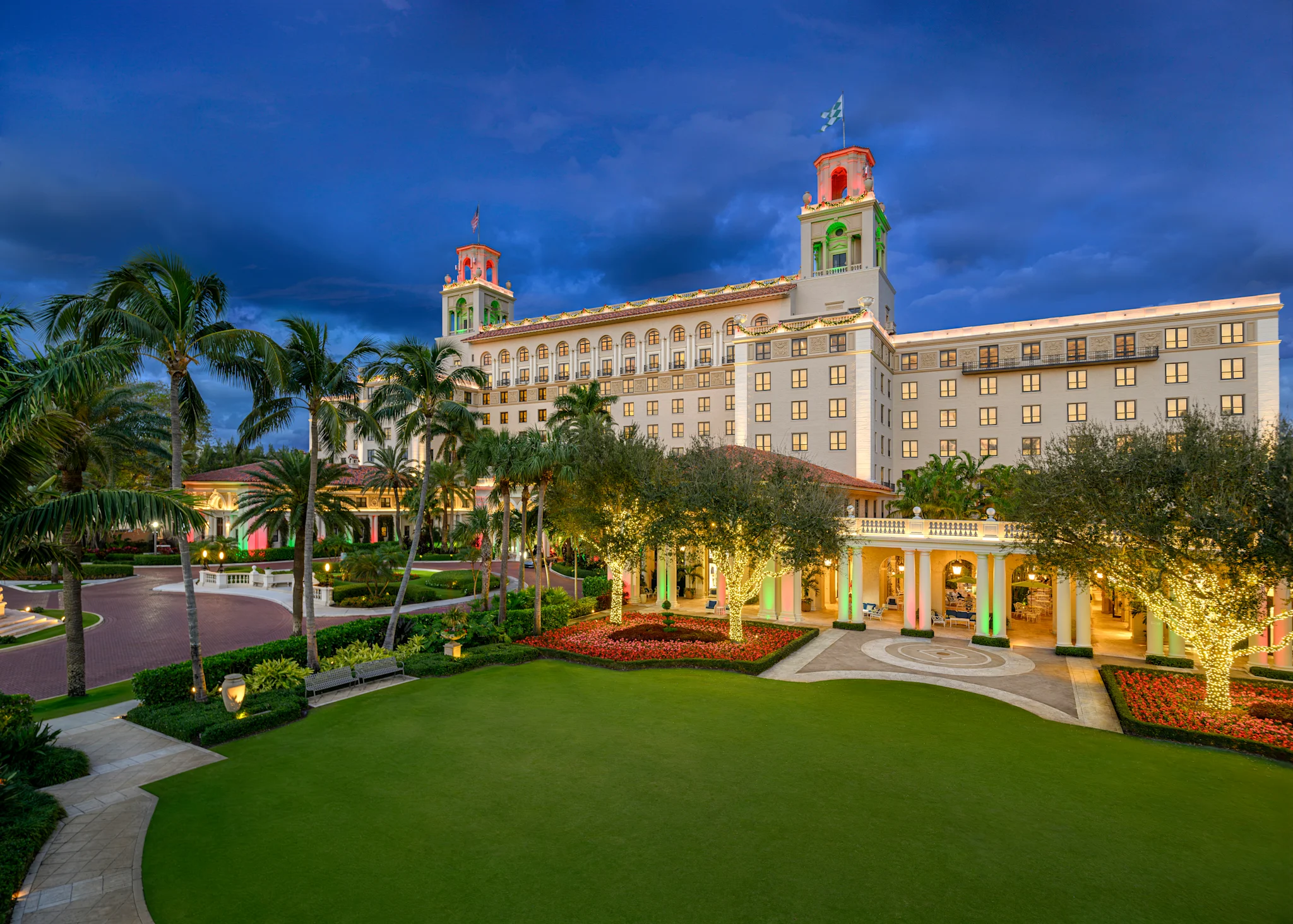 Illuminated luxury hotel with twin towers at dusk, surrounded by palm trees and manicured lawns.