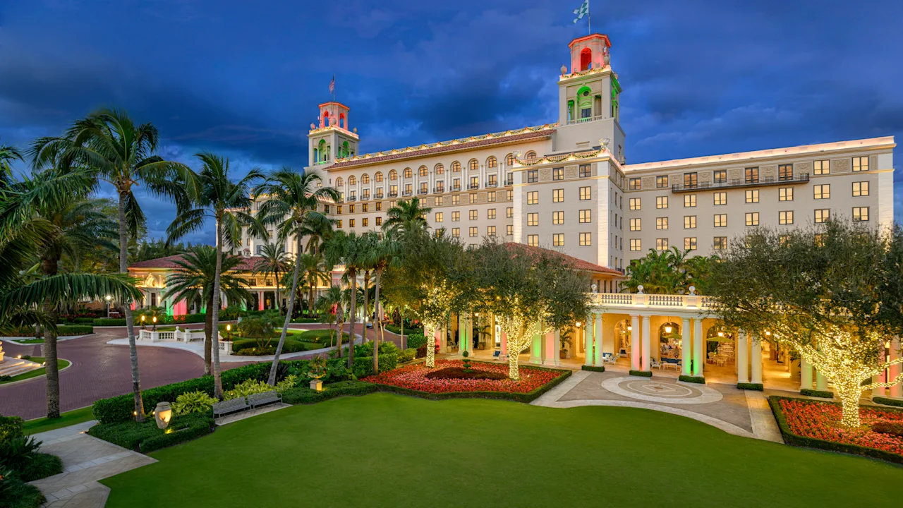 Illuminated luxury hotel with twin towers at dusk, surrounded by palm trees and manicured lawns.