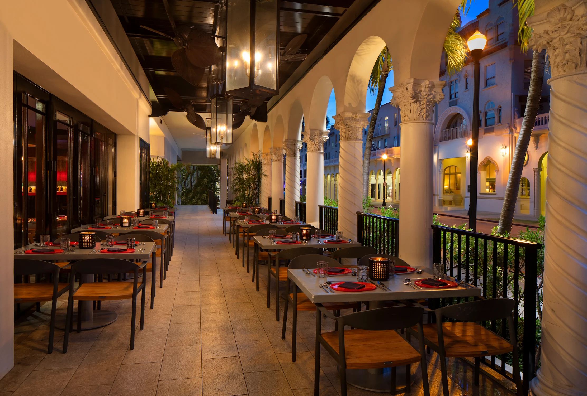 Elegant outdoor restaurant terrace with white columns, dark wood tables set with red placemats at dusk.