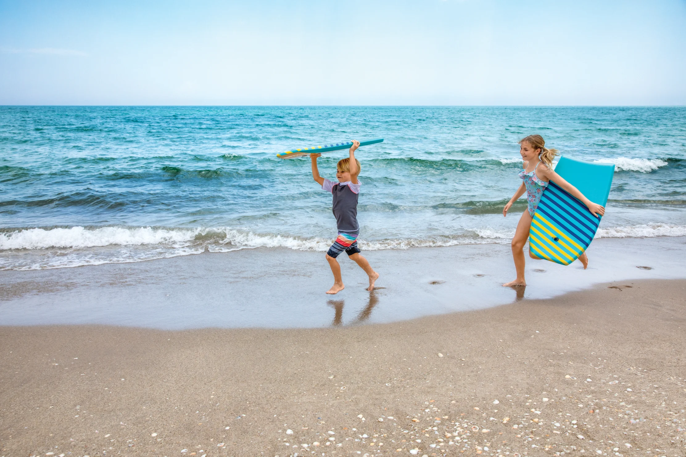 Two children running on a sandy beach with colorful striped boogie boards near turquoise ocean water.