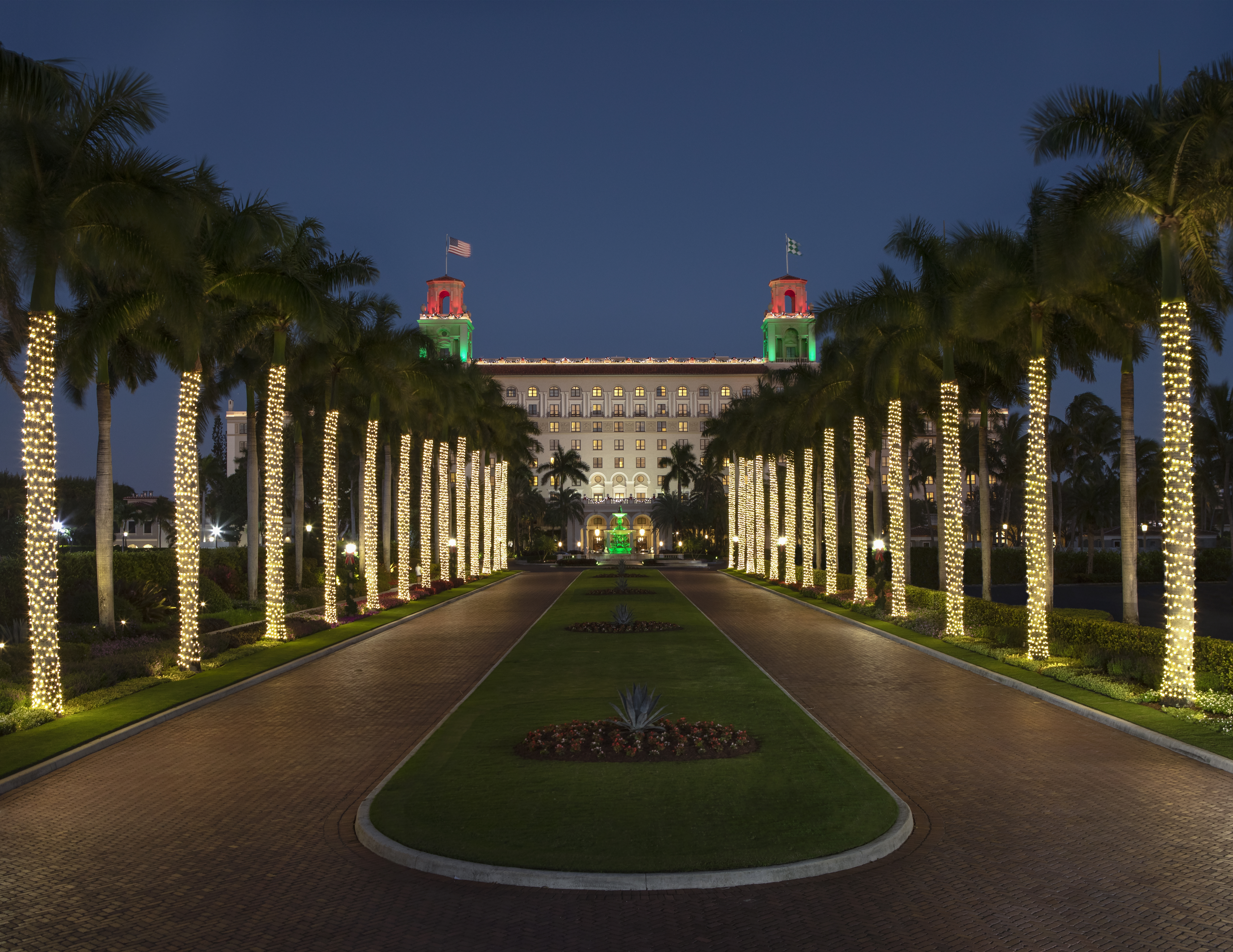 Illuminated palm trees line a driveway leading to a grand hotel with red and green lit towers at dusk.