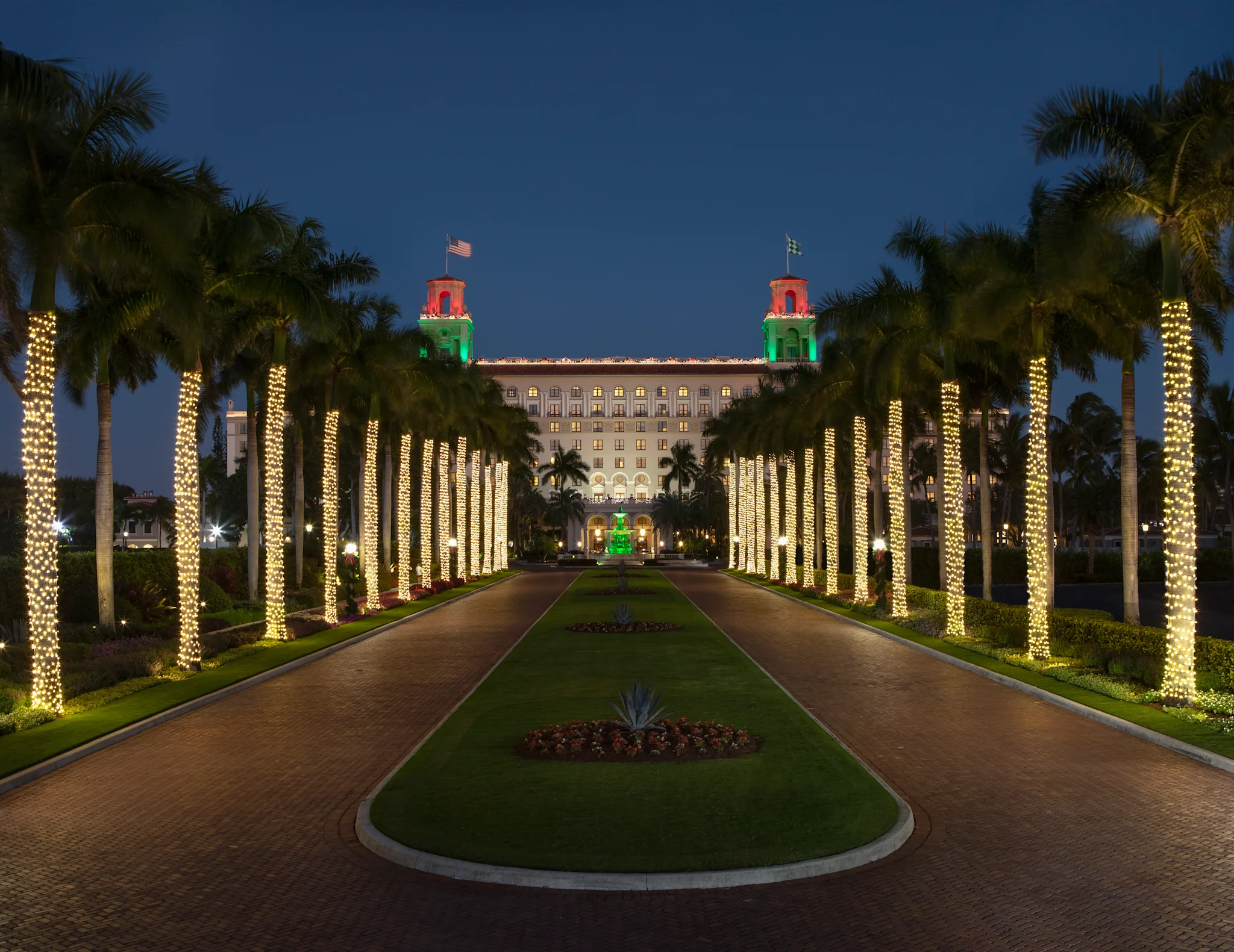 Illuminated palm trees line a driveway leading to a grand hotel with red and green lit towers at dusk.