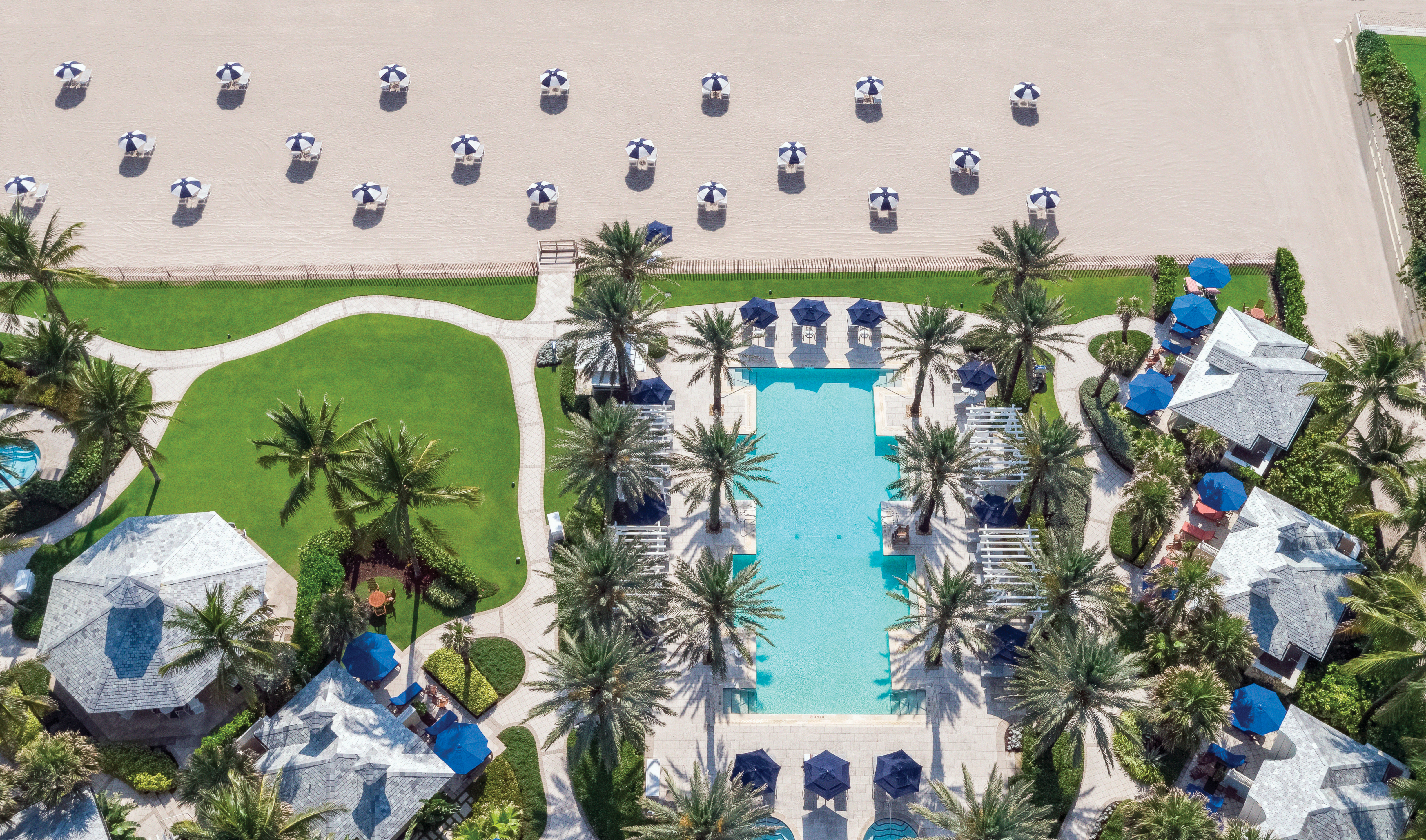 Aerial view of a luxury beach resort with turquoise pool, palm trees, cabanas, and striped umbrellas on sand.