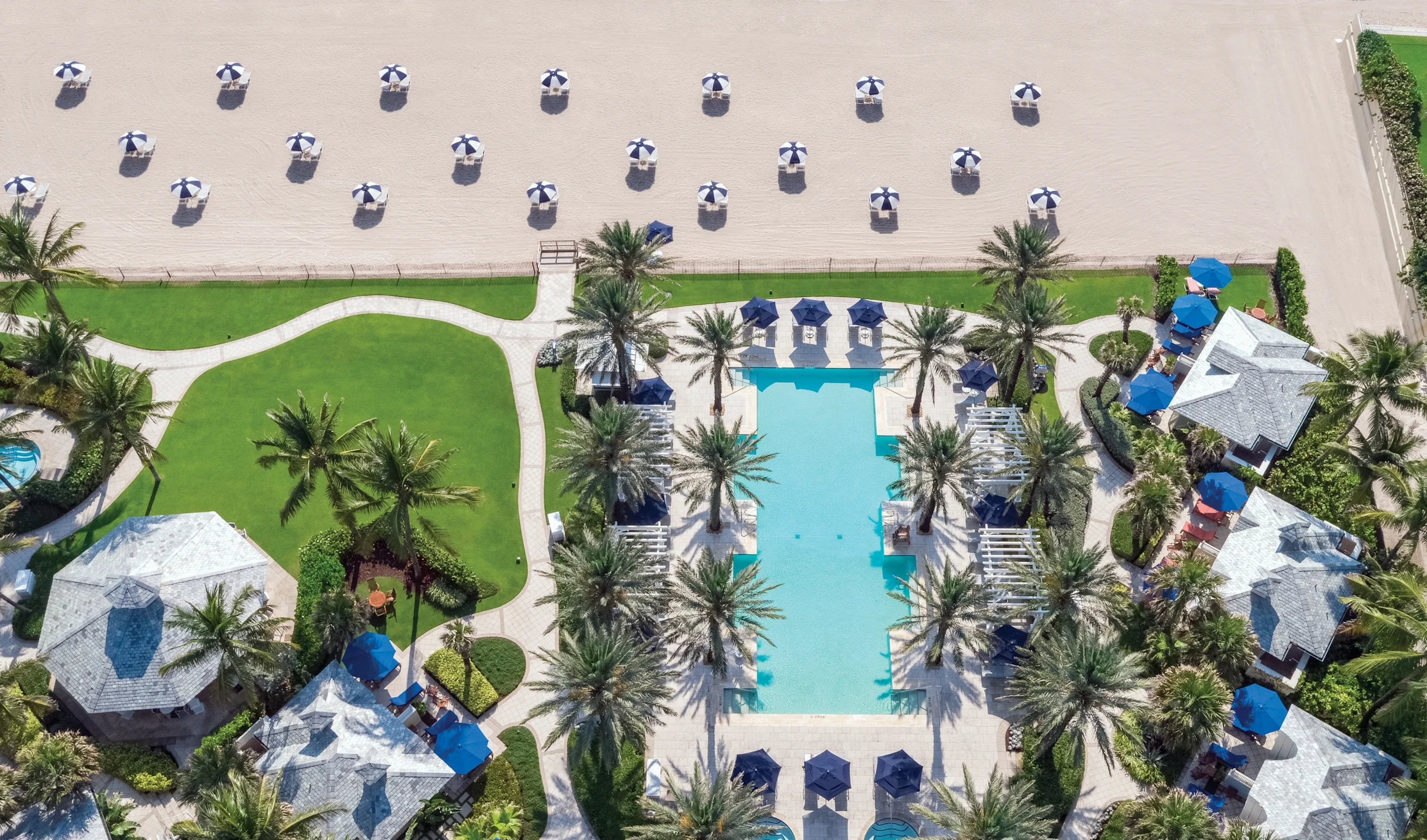 Aerial view of a luxury beach resort with turquoise pool, palm trees, cabanas, and striped umbrellas on sand.