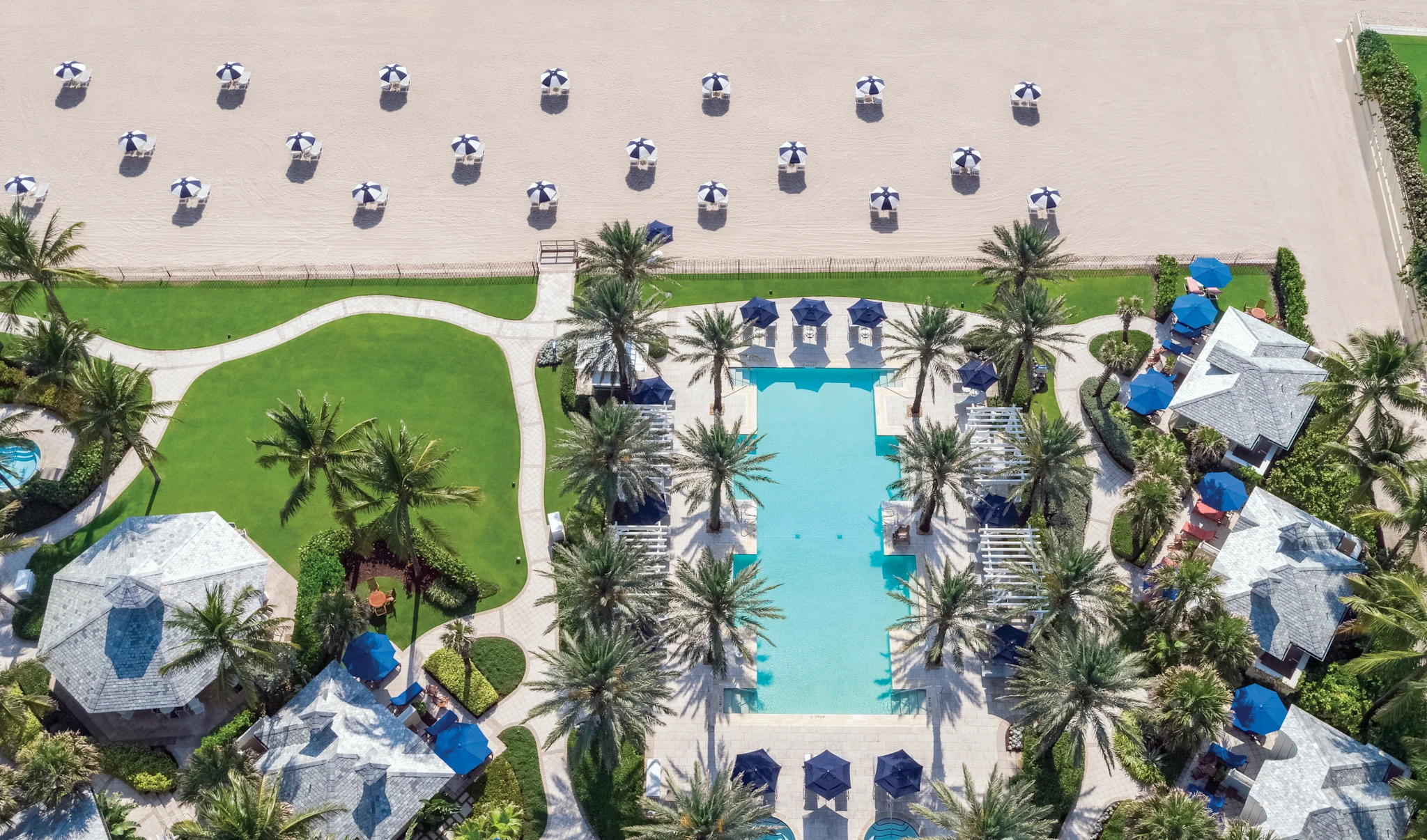 Aerial view of a luxury beach resort with turquoise pool, palm trees, cabanas, and striped umbrellas on sand.