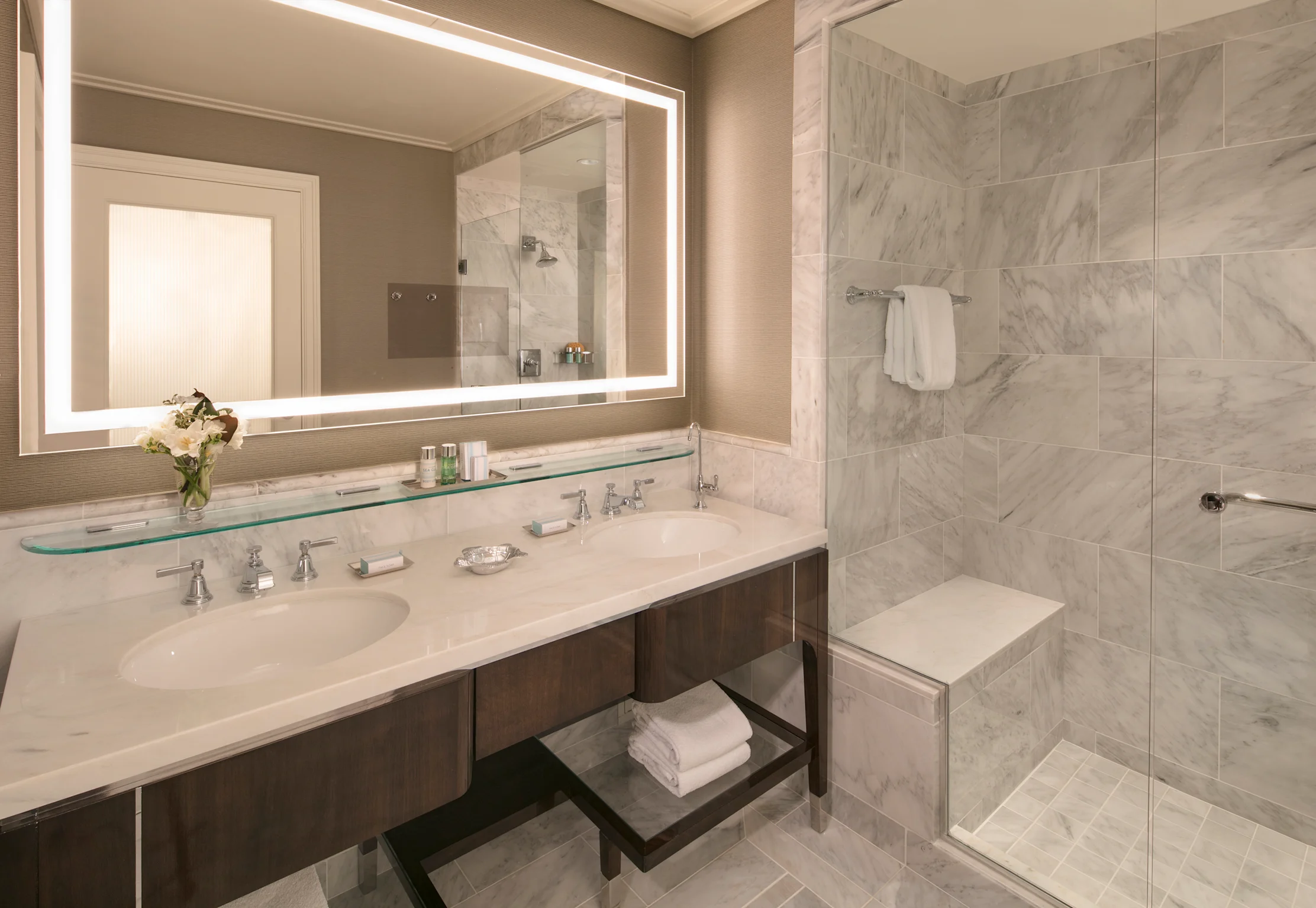 Modern hotel bathroom with marble tiles, double vanity, backlit mirror, and glass-enclosed shower.