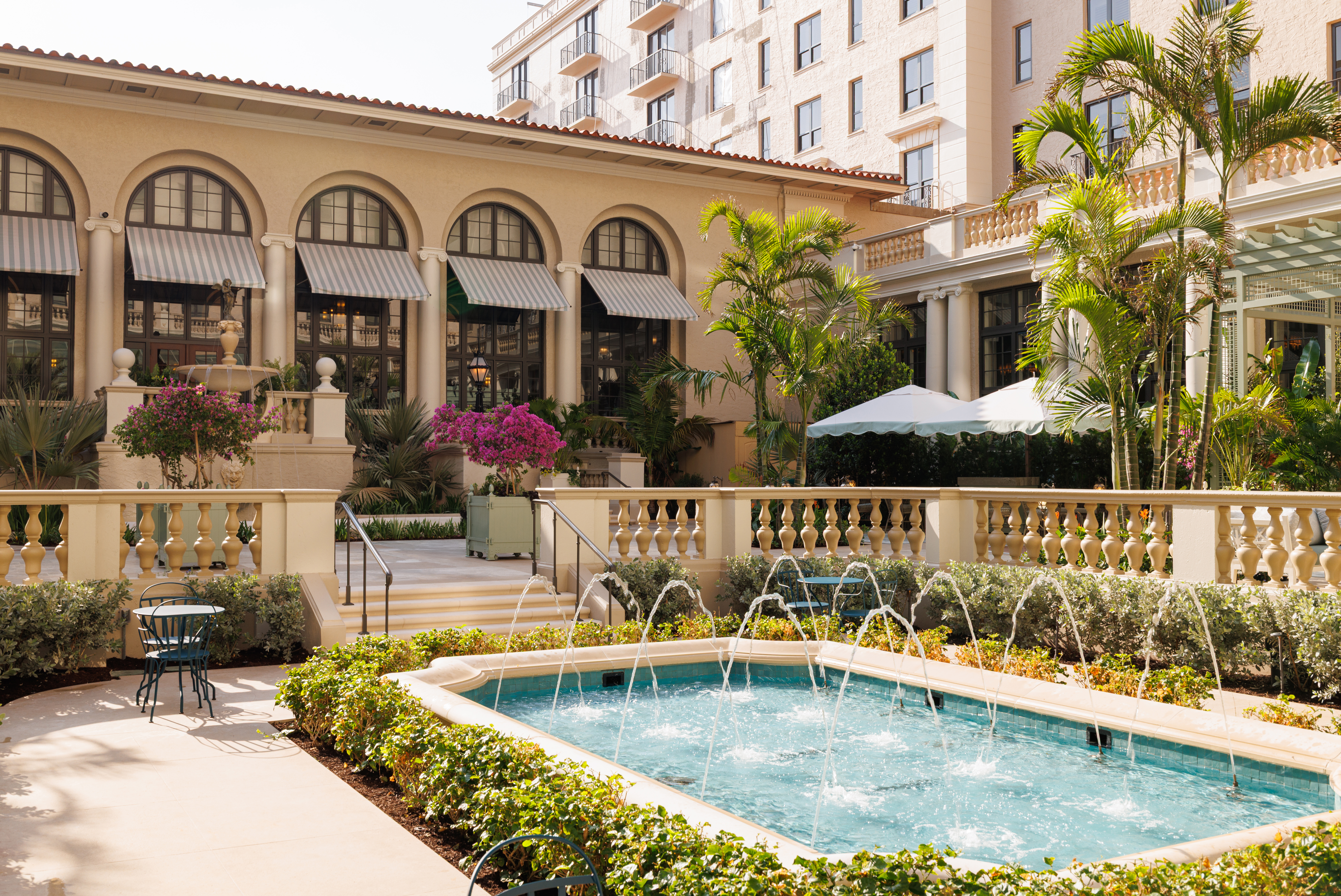 Mediterranean-style hotel courtyard with fountain, palm trees, arched colonnade, and beige buildings.