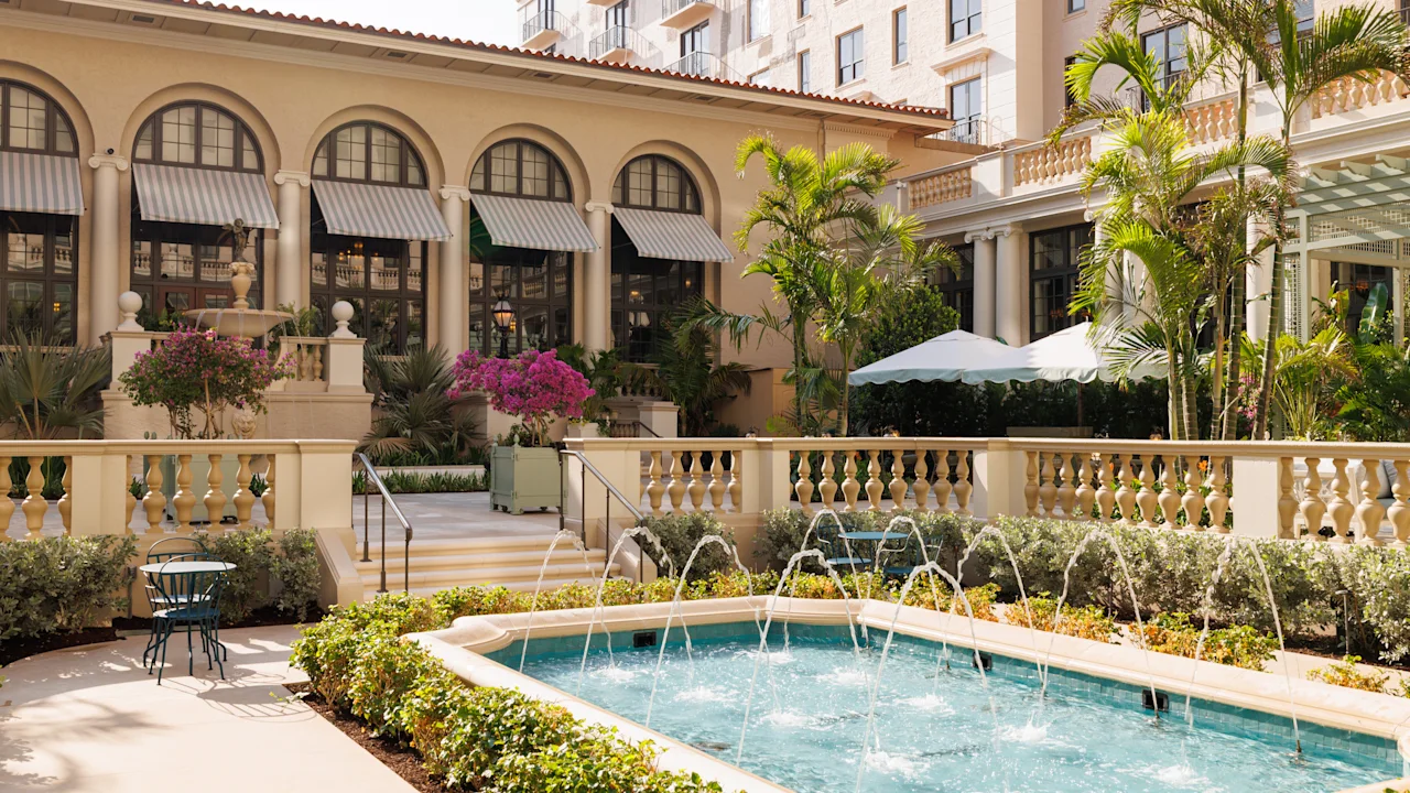 Mediterranean-style hotel courtyard with fountain, palm trees, arched colonnade, and beige buildings.