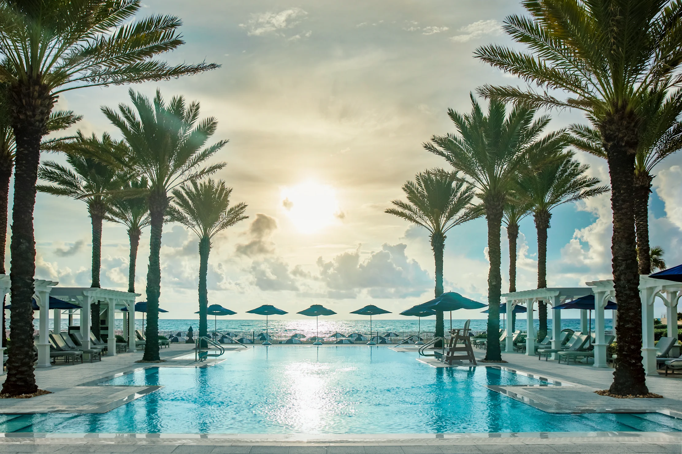 Tropical resort pool framed by palm trees with ocean view, white cabanas, and dramatic cloudy sky.