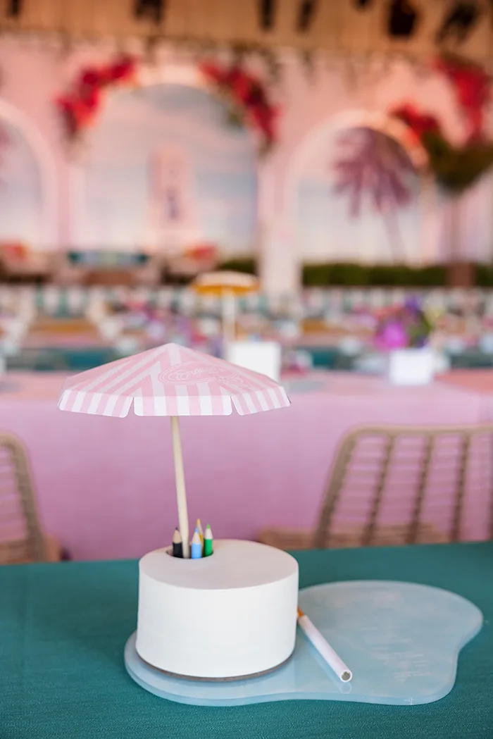 White circular notepad with pink striped umbrella topper on blue pool-shaped plate with colored pencils at party table setting