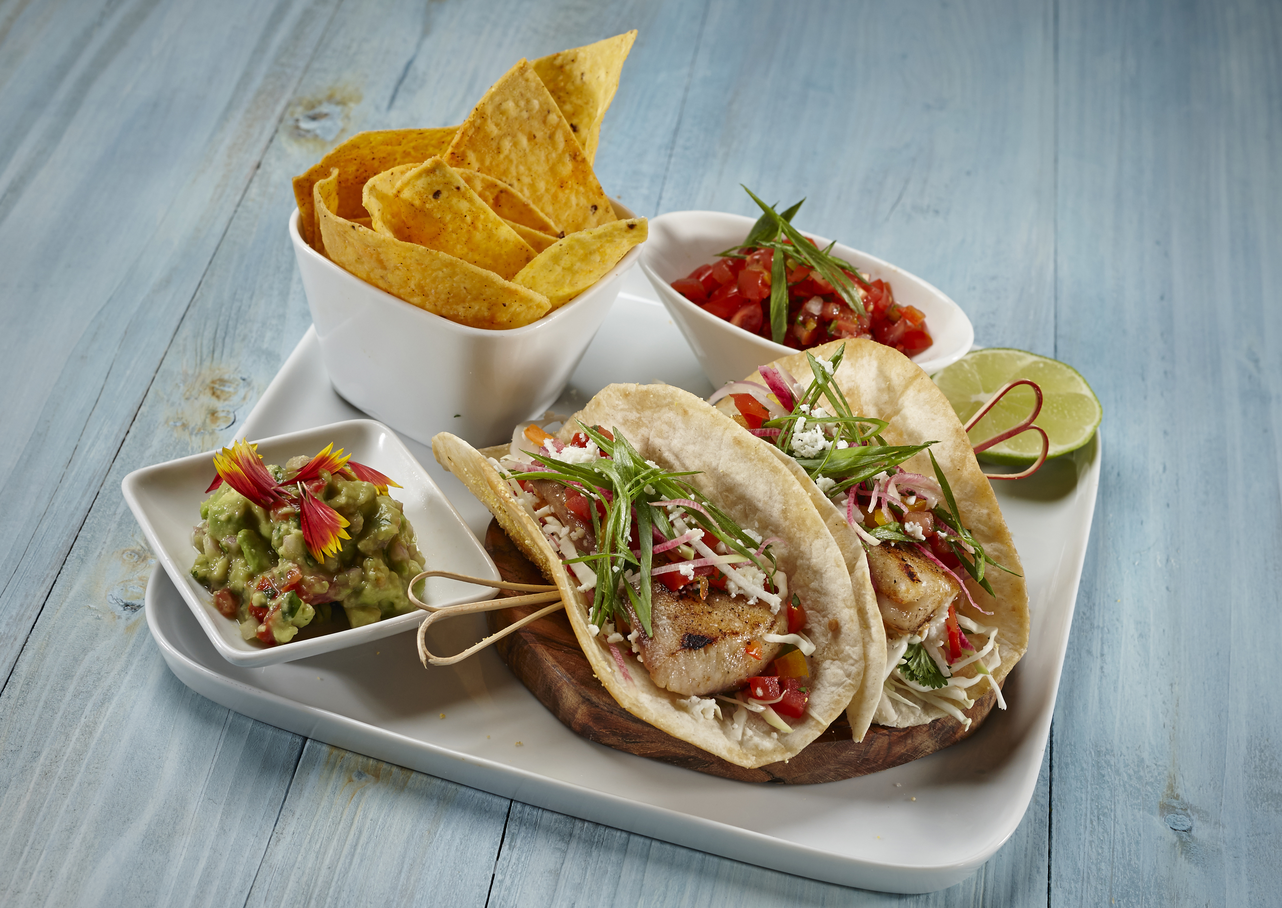 Fish tacos with cabbage slaw, salsa, guacamole, and tortilla chips on a white platter with lime wedge.