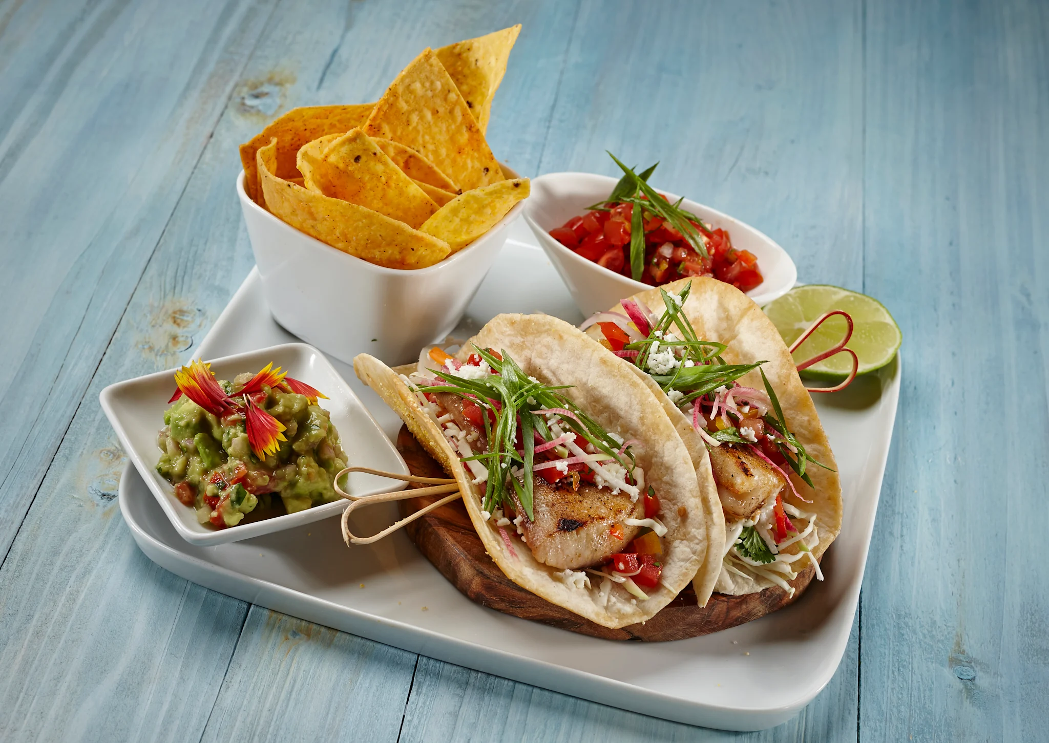 Fish tacos with cabbage slaw, salsa, guacamole, and tortilla chips on a white platter with lime wedge.