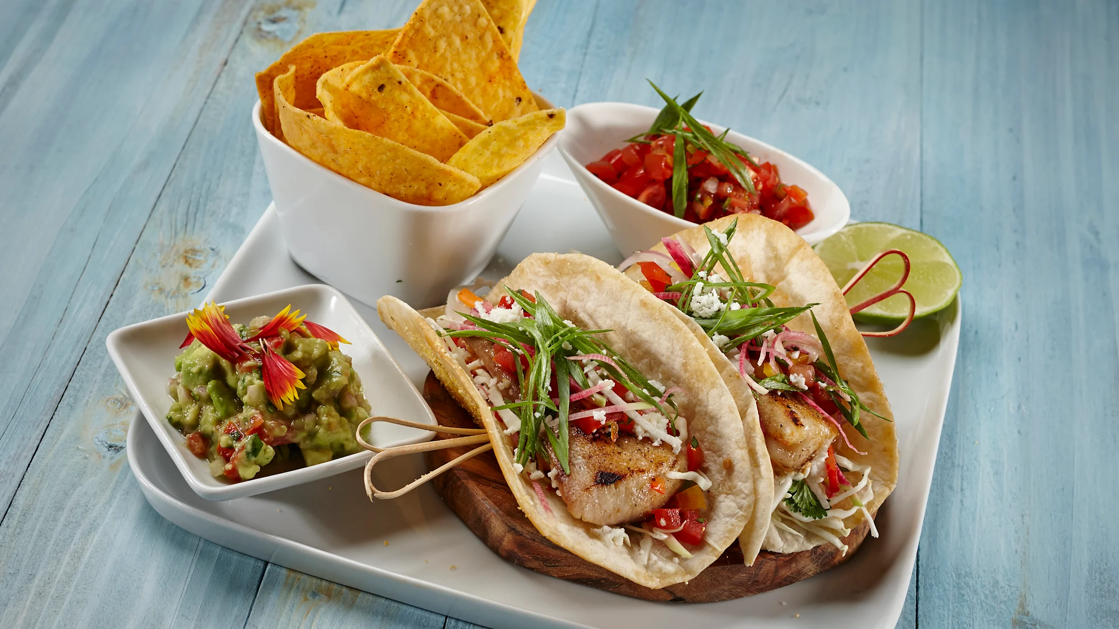 Fish tacos with cabbage slaw, salsa, guacamole, and tortilla chips on a white platter with lime wedge.