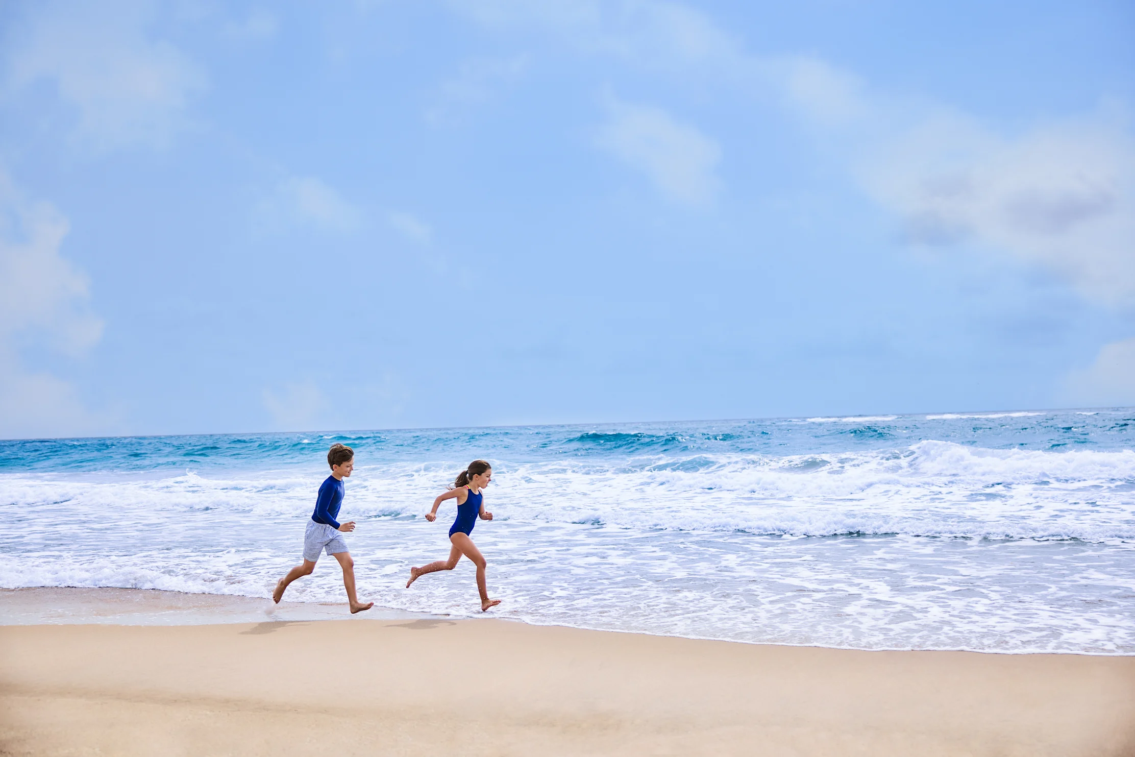 Two children in blue swimwear running along a sandy beach with turquoise waves and blue sky.