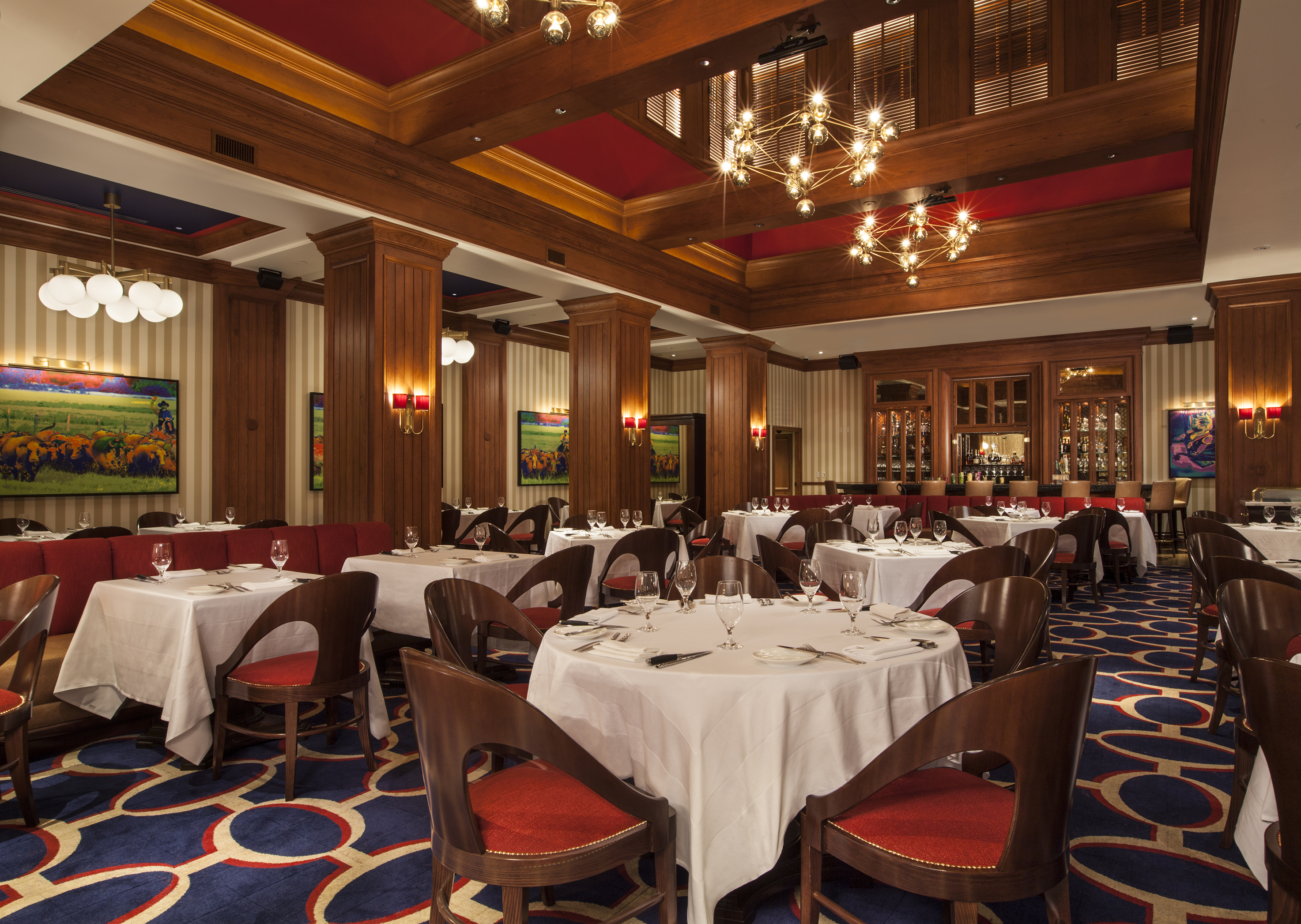 Upscale restaurant interior with white-clothed tables, red velvet chairs, wood paneling, and chandeliers.