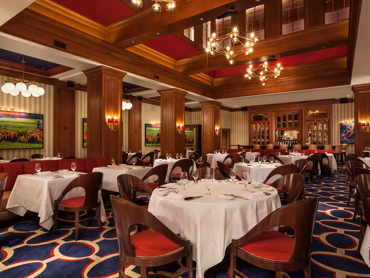 Upscale restaurant interior with white-clothed tables, red velvet chairs, wood paneling, and chandeliers.