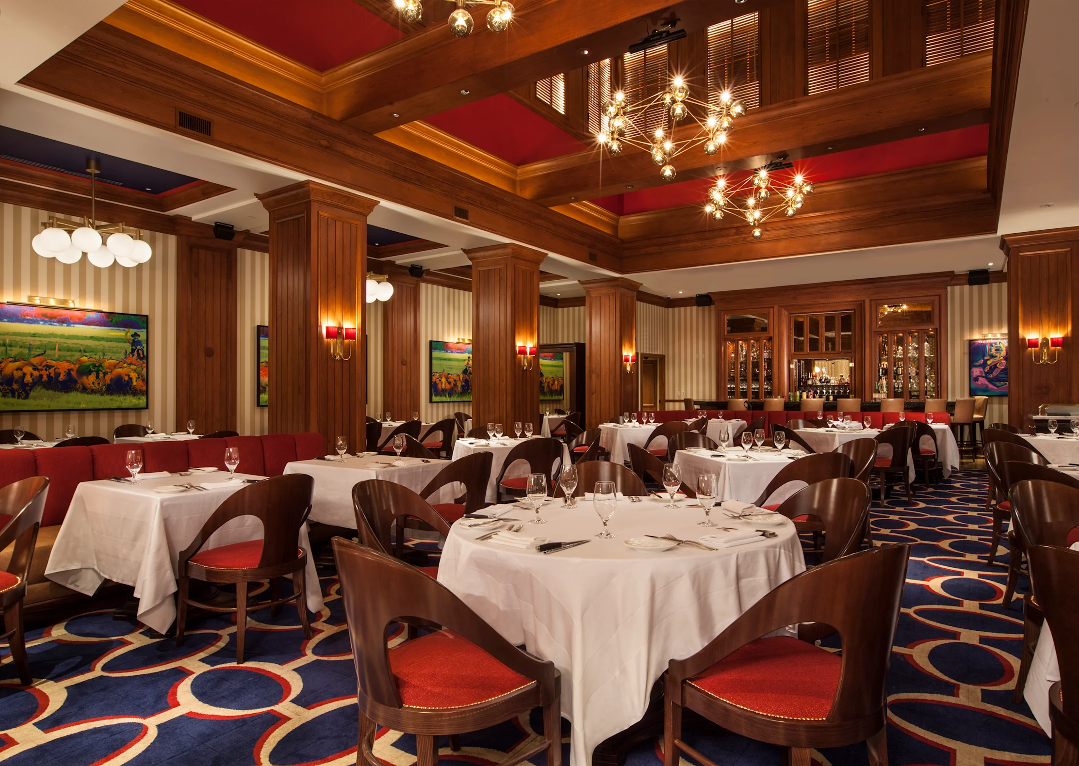 Upscale restaurant interior with white-clothed tables, red velvet chairs, wood paneling, and chandeliers.