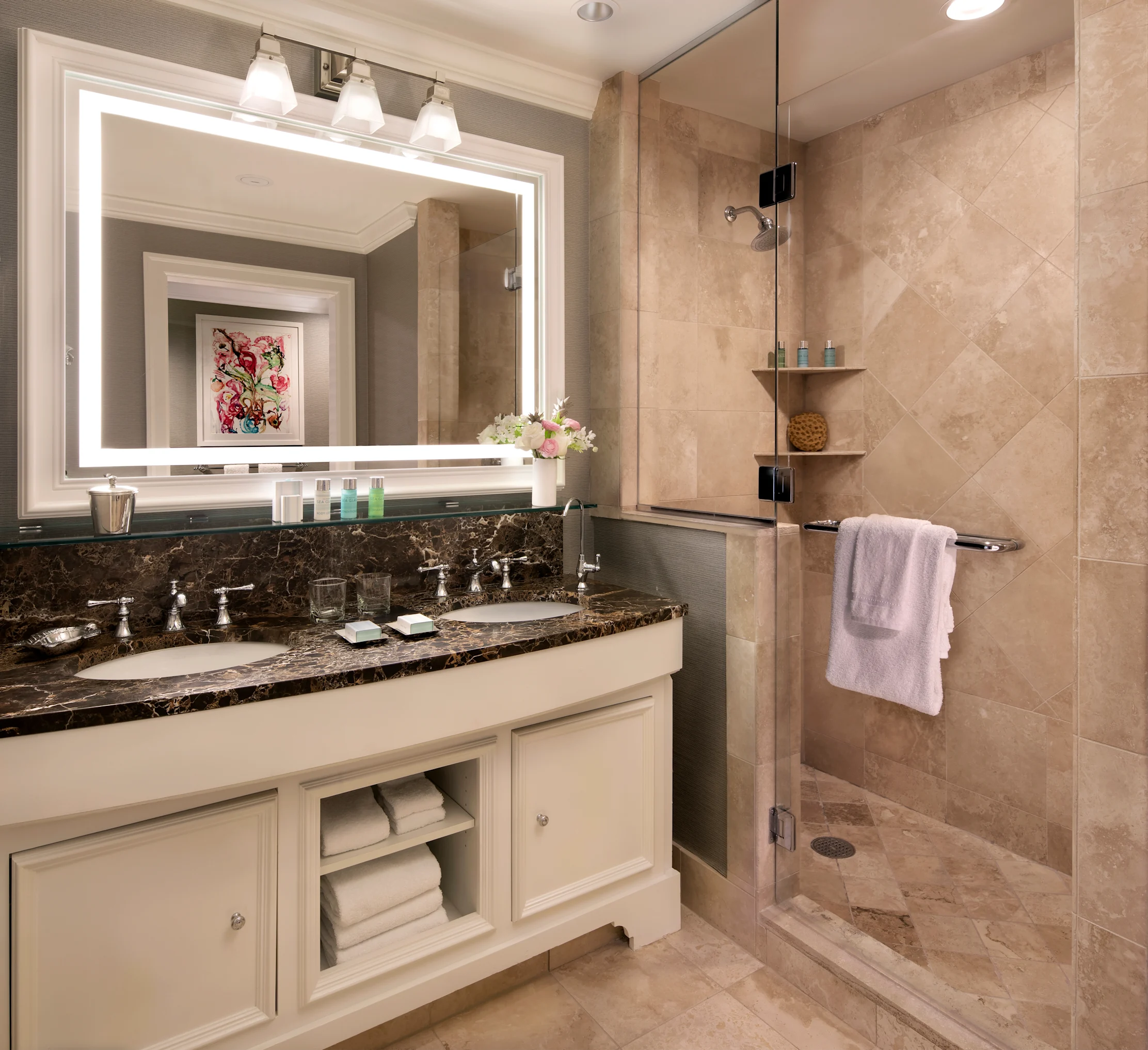 Modern bathroom with white vanity, dark marble countertop, lighted mirror, and beige tiled shower.