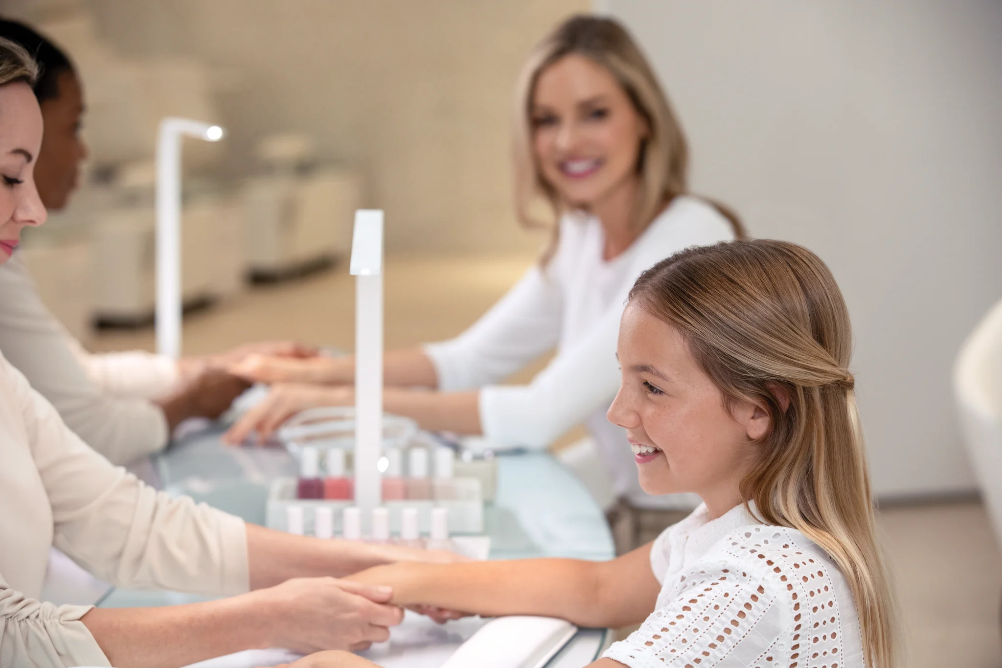 Young girl receiving a manicure at a salon with staff members assisting and smiling in background.
