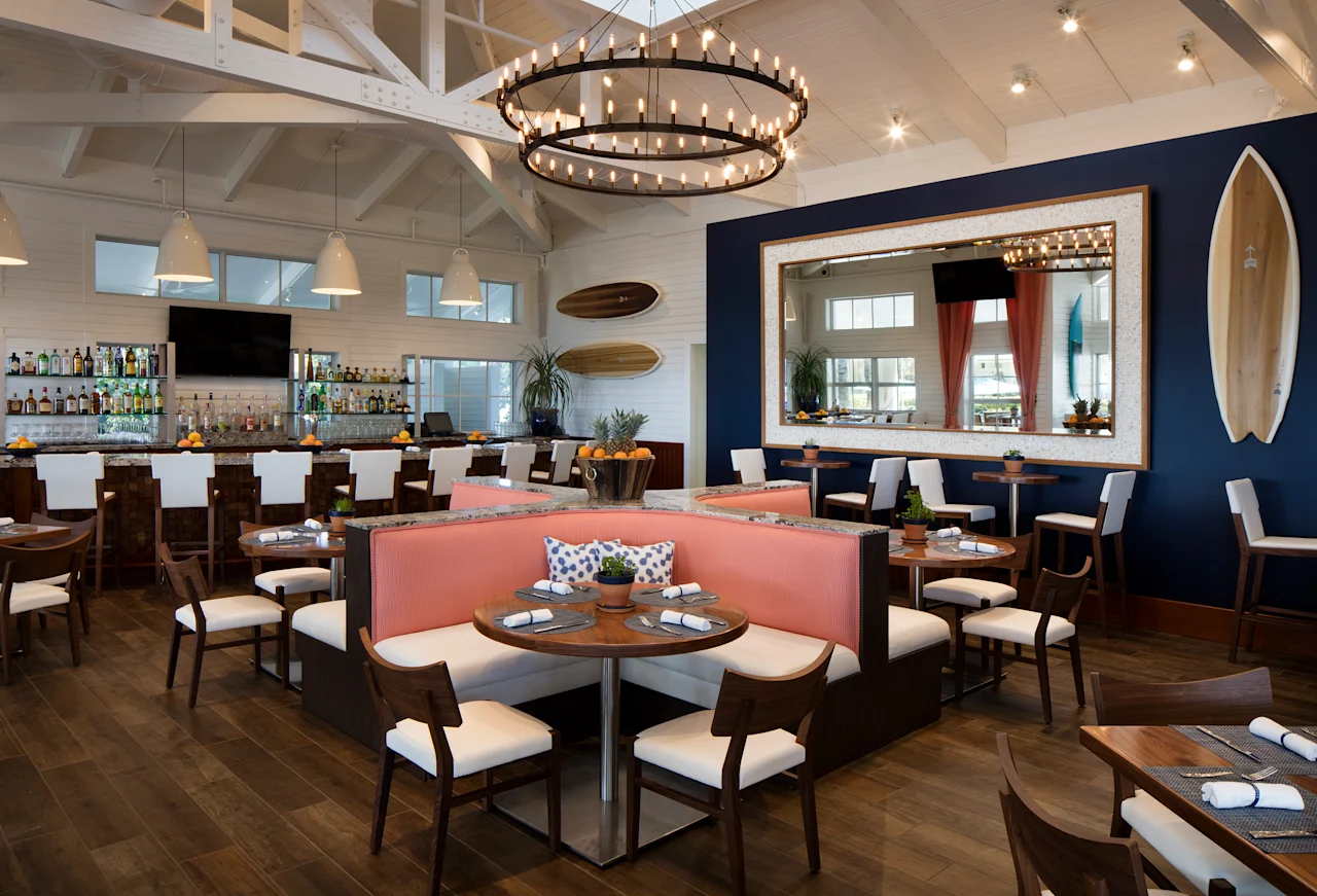 Modern coastal restaurant interior with white vaulted ceiling, circular chandelier, bar area, and coral banquette seating.