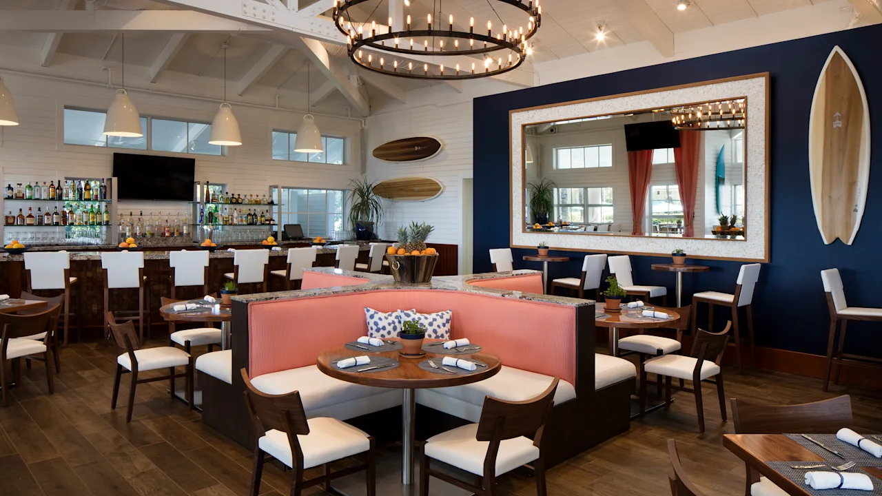 Modern coastal restaurant interior with white vaulted ceiling, circular chandelier, bar area, and coral banquette seating.