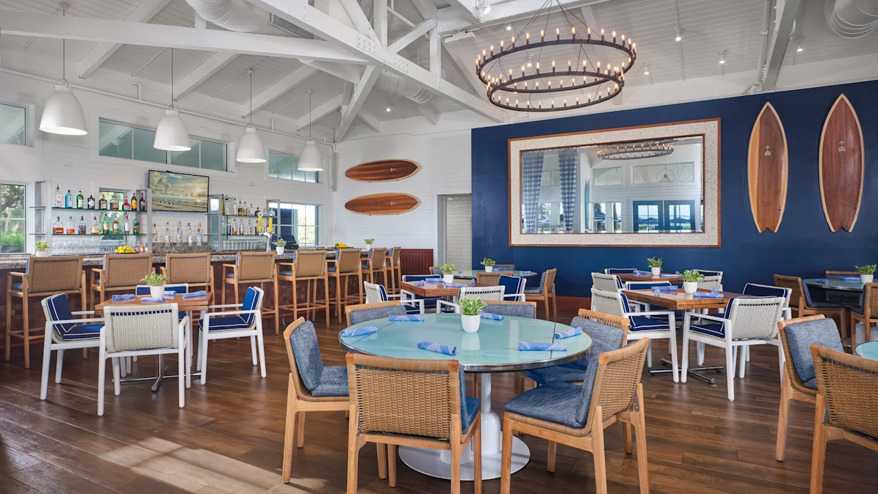 Modern coastal restaurant interior with white vaulted ceiling, circular chandelier, bar area, and coral banquette seating.
