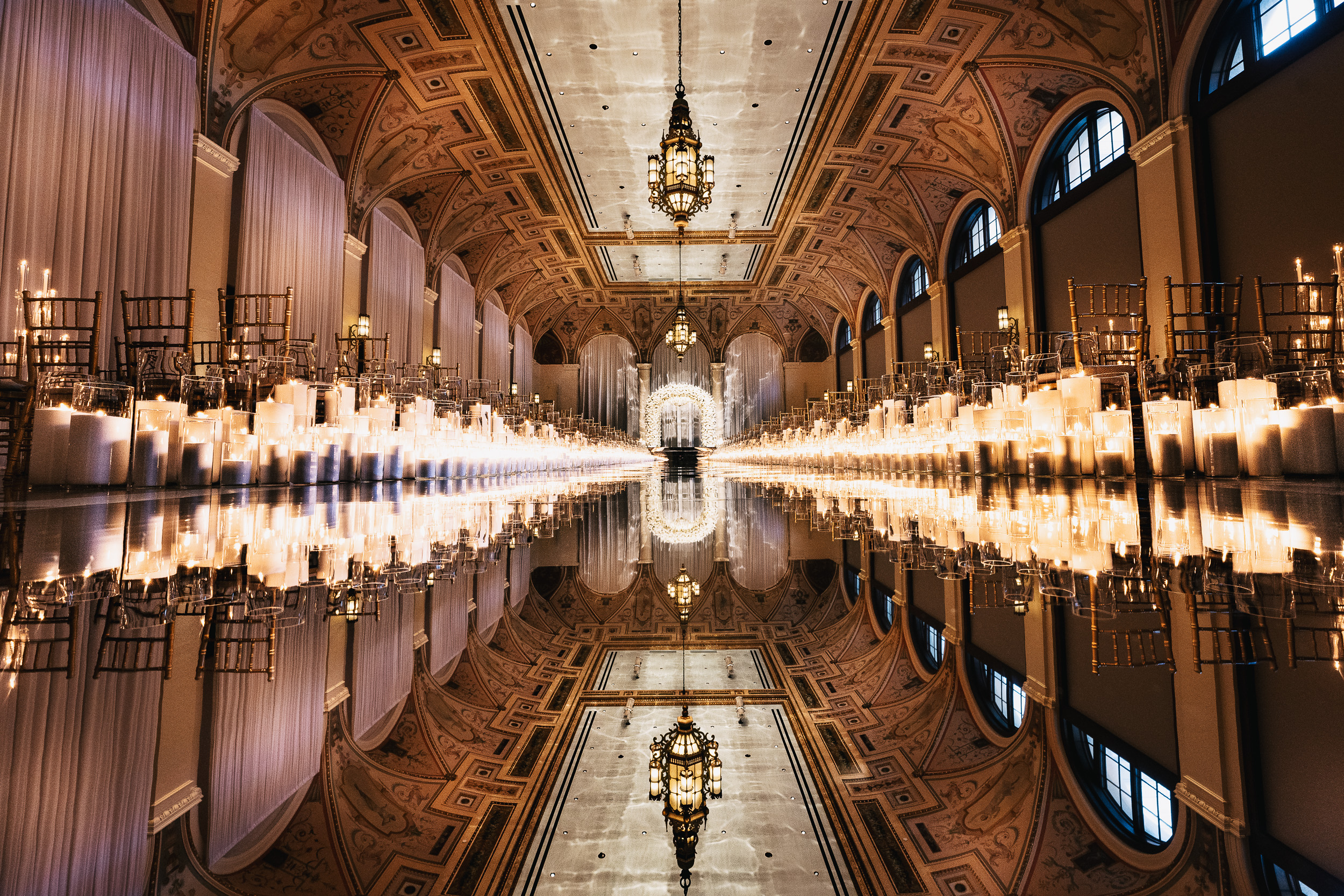 Grand ornate hall with vaulted ceilings, rows of candles lining a reflective aisle, and gold chairs.