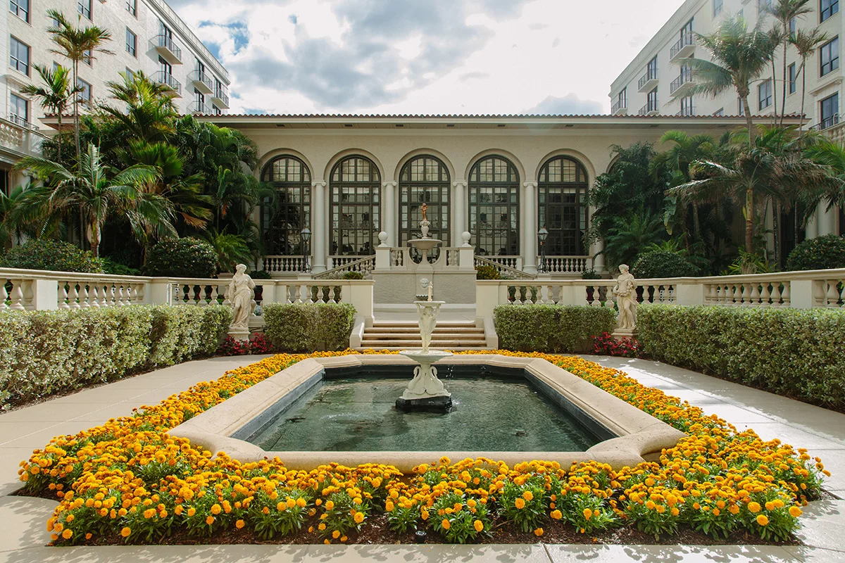 Mediterranean-style courtyard with fountain surrounded by yellow flowers, palm trees, and arched colonnade.
