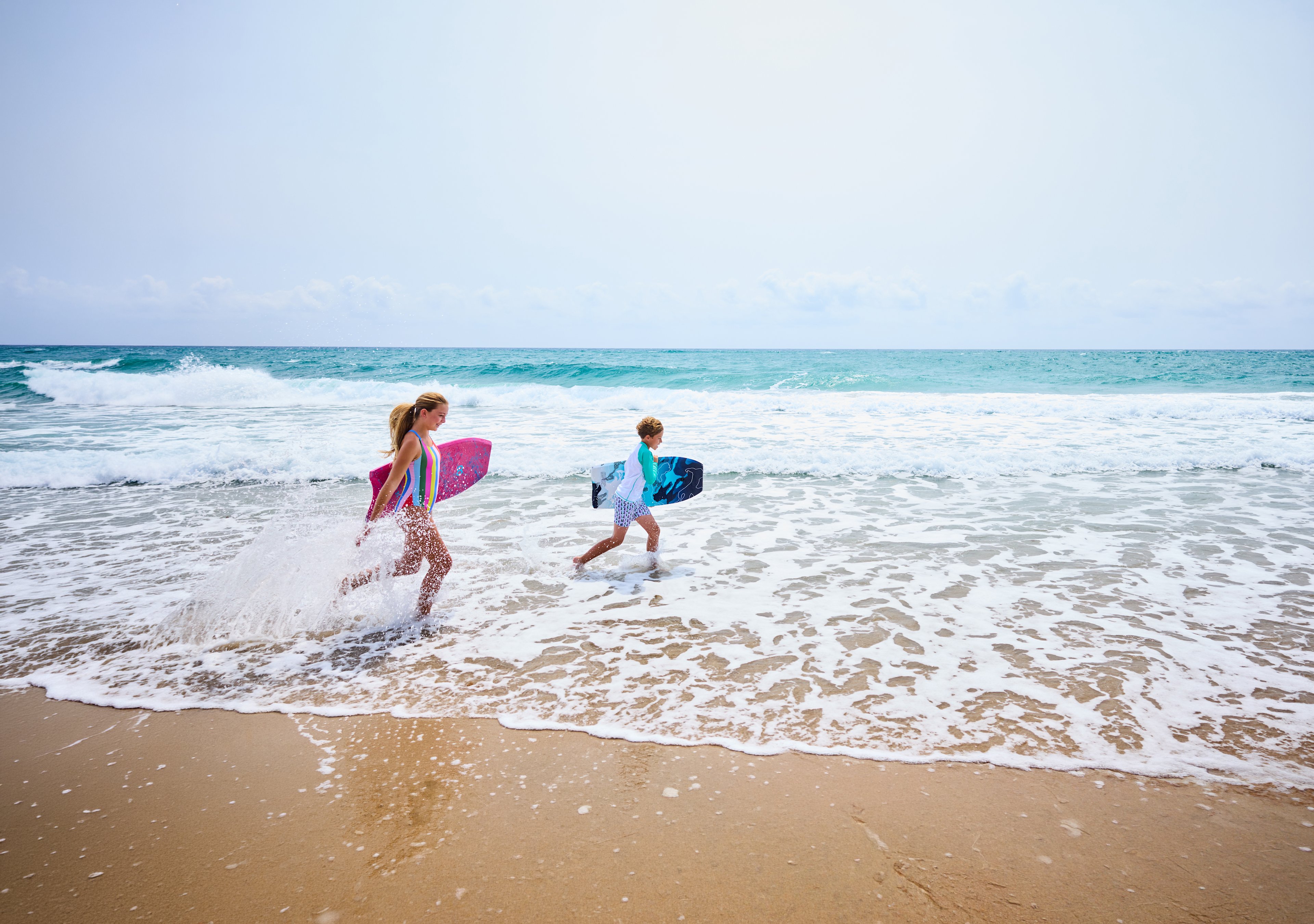 Two children running through ocean waves carrying colorful boogie boards on a sunny beach.