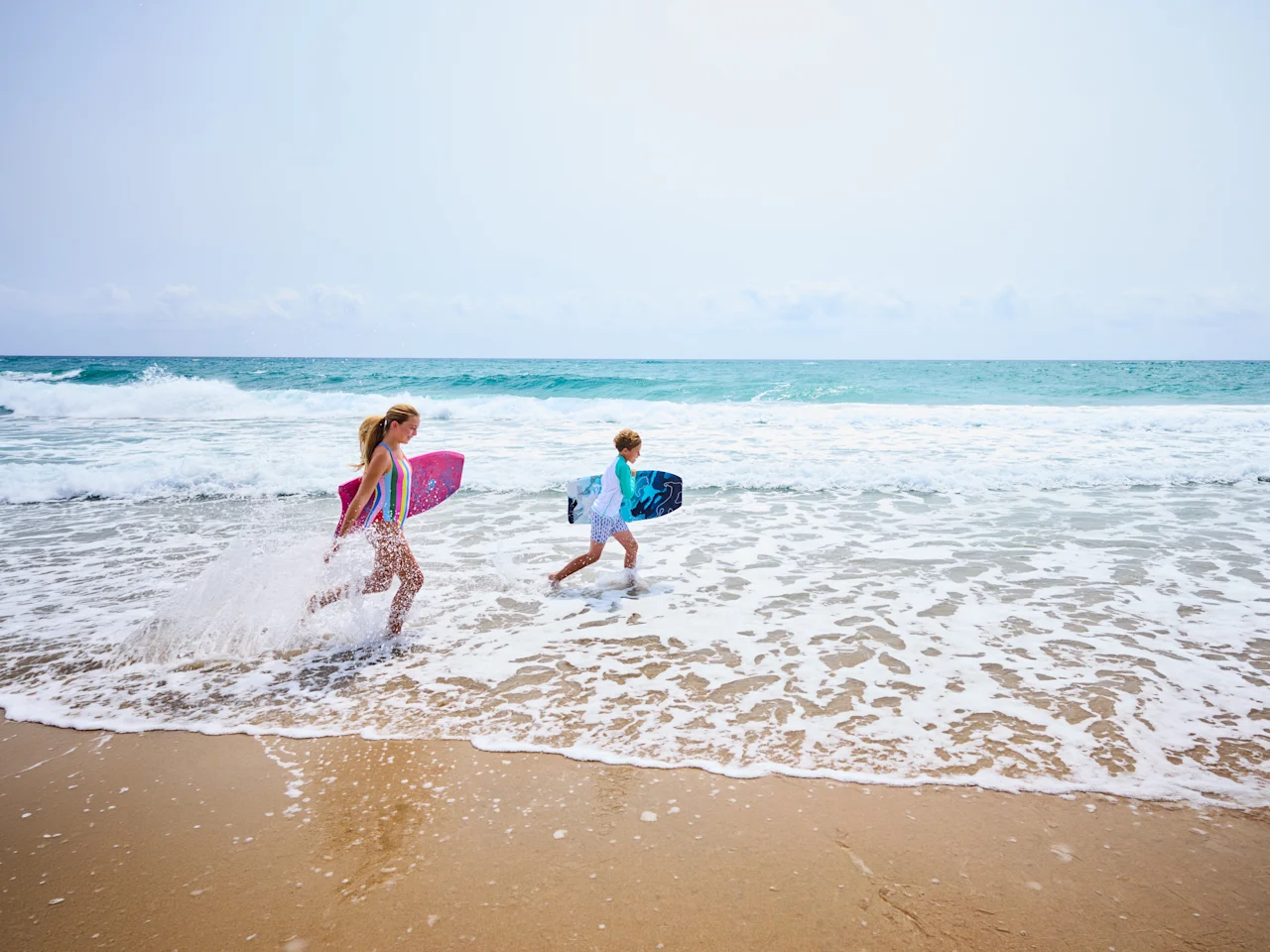 Two children running through ocean waves carrying colorful boogie boards on a sunny beach.