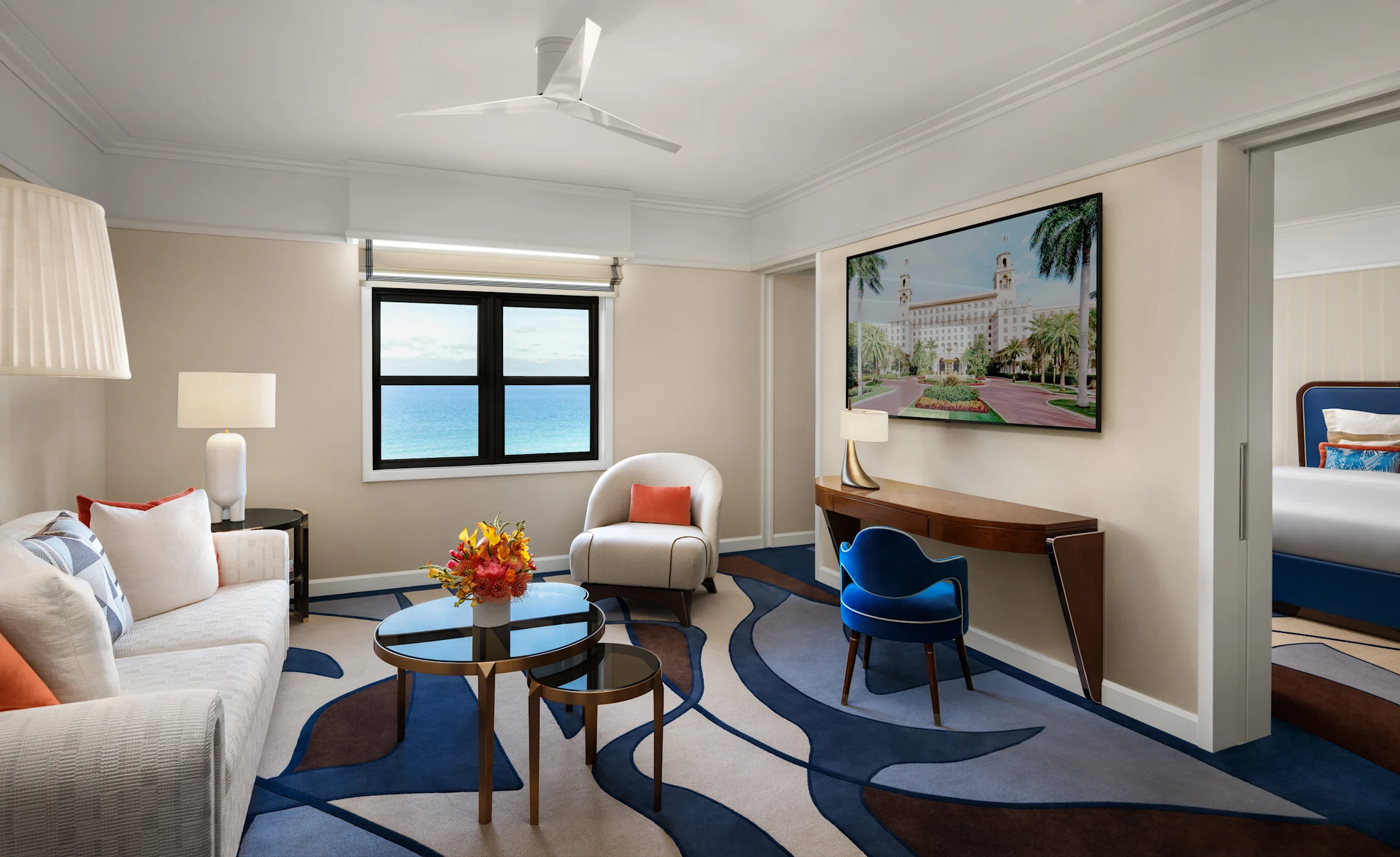 Modern hotel suite with ocean view window, beige sofa, blue accent chair, and framed artwork.