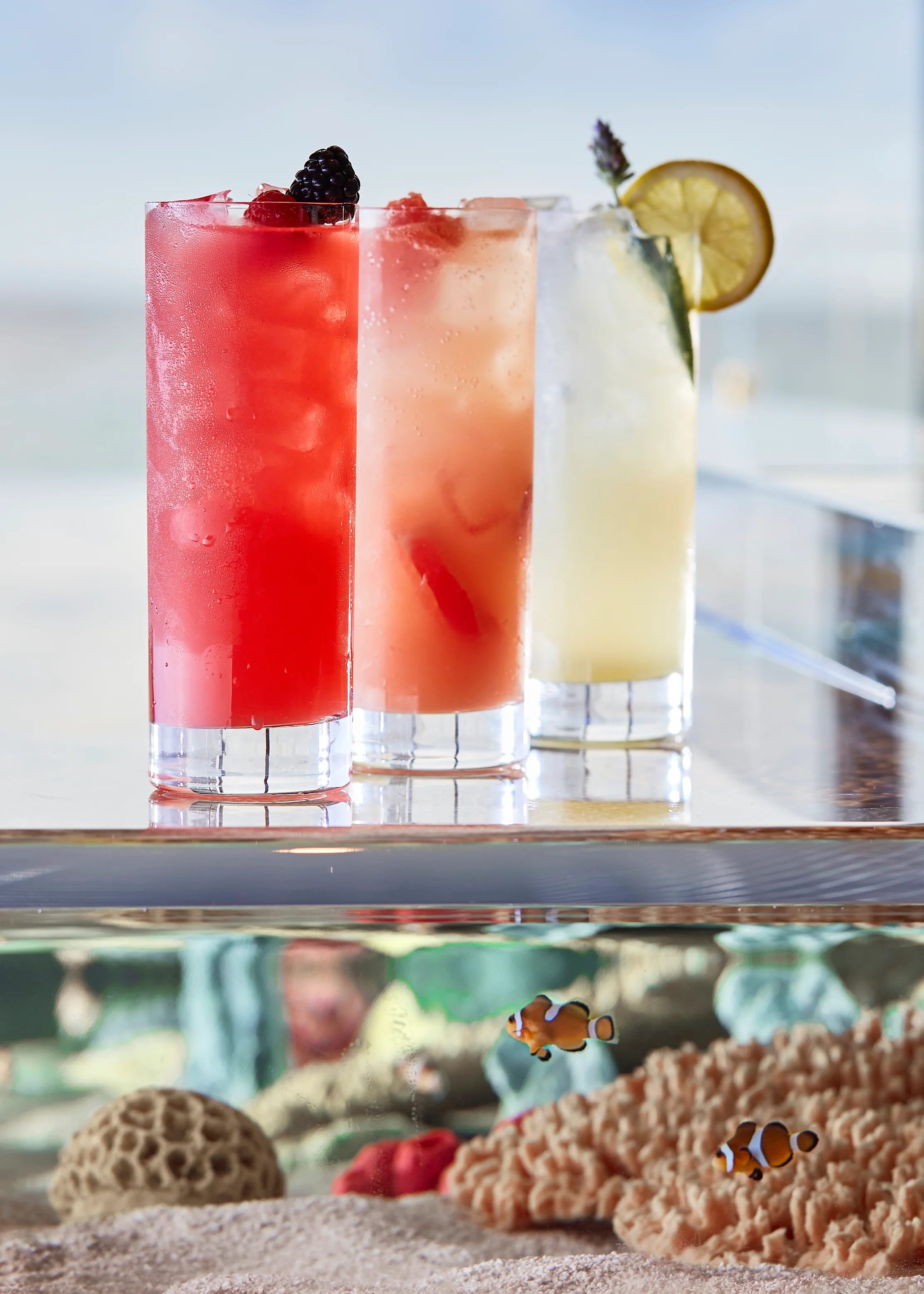 Three colorful cocktails with garnishes displayed on a glass surface above an aquarium with fish.