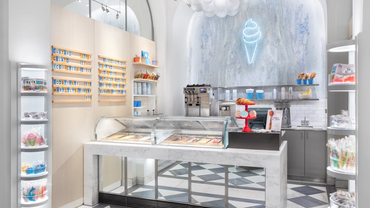 Modern ice cream shop interior with white and blue decor, neon cone sign, menu boards, and display cases.