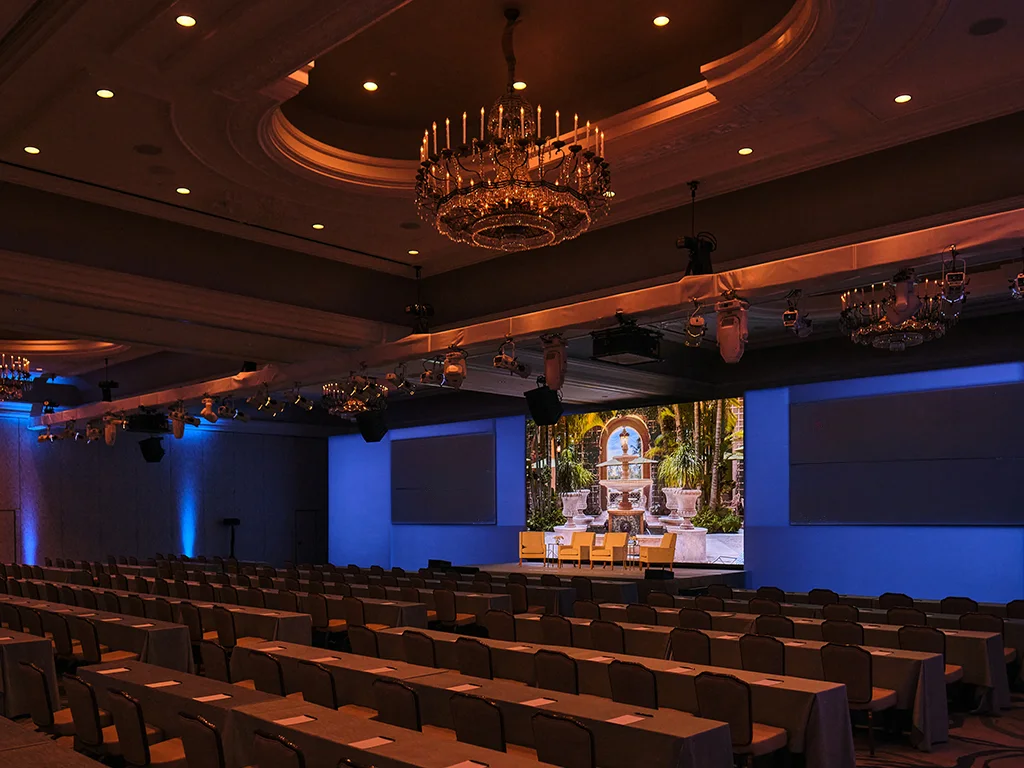 Ballroom set for a presentation with a center screen showing a courtyard fountain flanked by two blank side screens, rigged lighting, and rows of skirted tables beneath a crystal chandelier.
