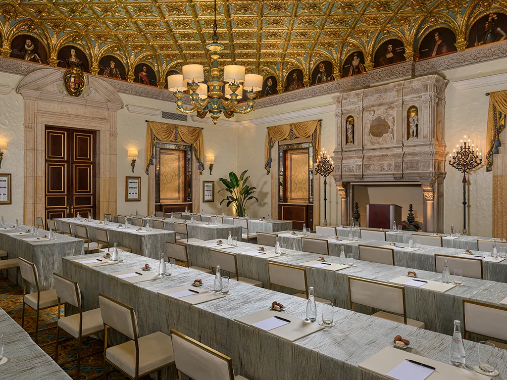 Meeting room set classroom-style with rows of skirted tables, water service and notepads, beneath a gilded coffered ceiling lined with portrait paintings and a carved stone fireplace.