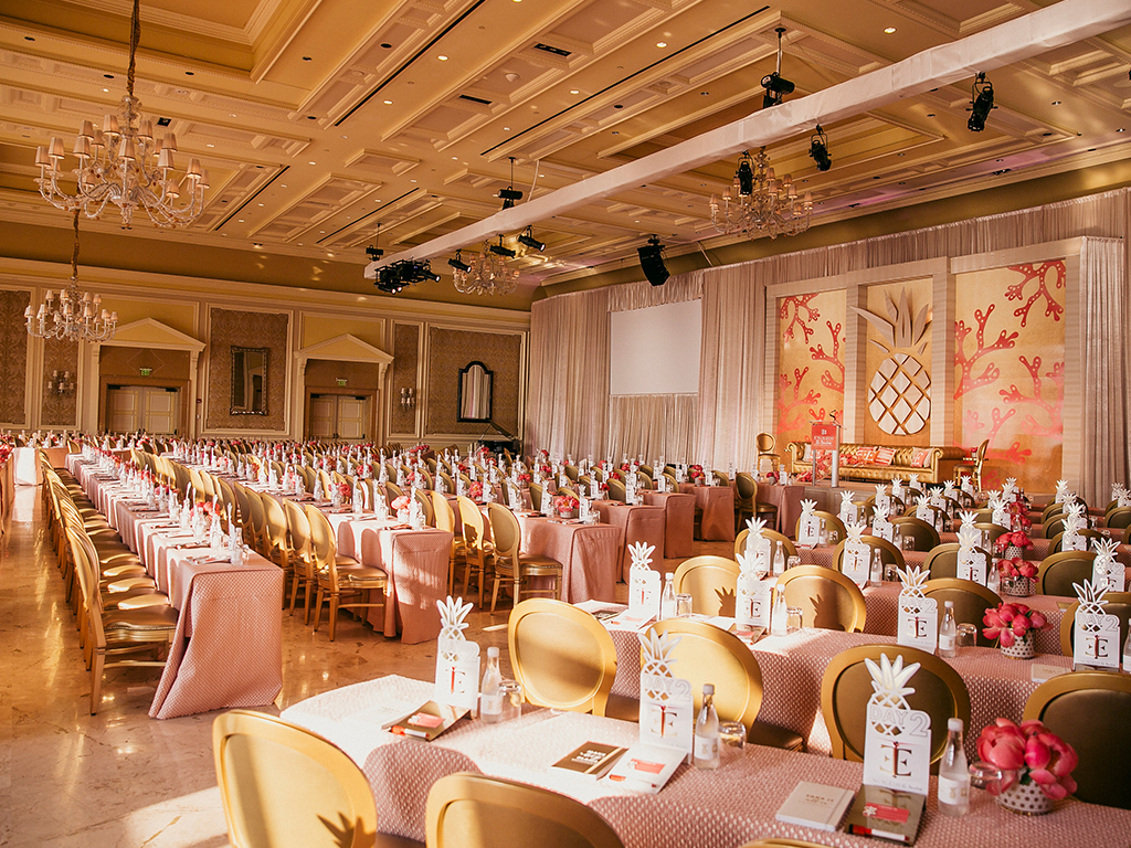 Conference room with rigged lighting truss and a projection screen above a stage with coral and pineapple motif panels, rows of pink-linened tables set with pineapple place-card holders.