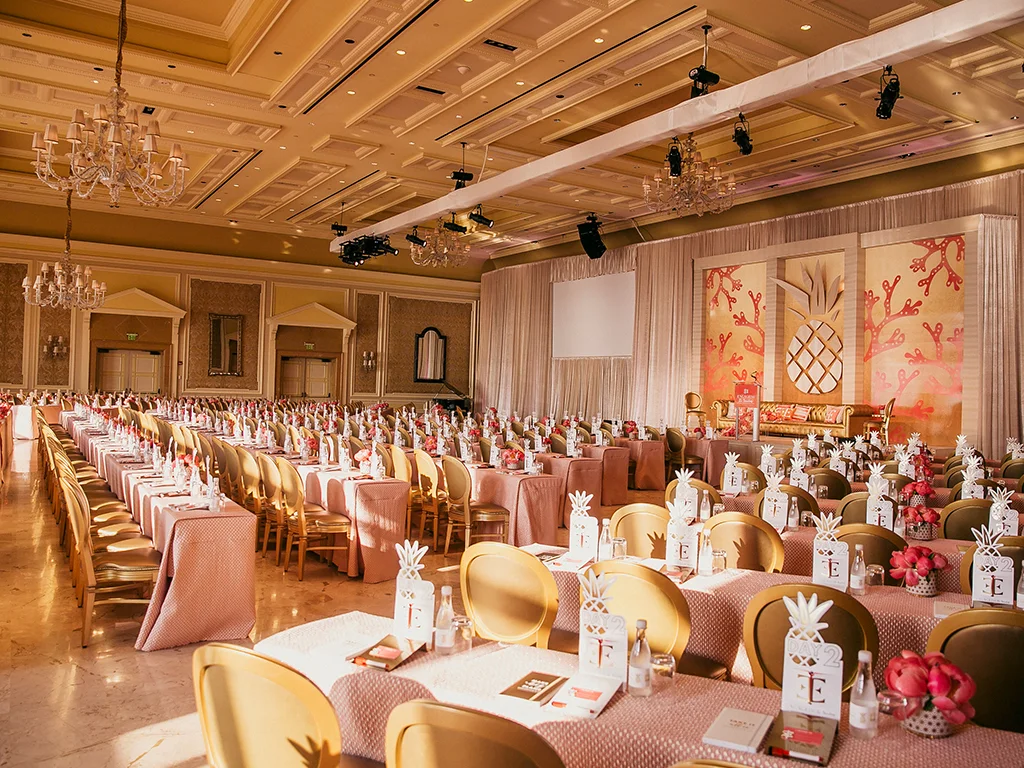 Conference room with rigged lighting truss and a projection screen above a stage with coral and pineapple motif panels, rows of pink-linened tables set with pineapple place-card holders.
