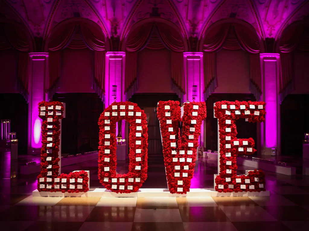 Oversized freestanding LOVE letters covered in red roses and studded with white seating cards, lit magenta beneath a colonnaded, arched ballroom ceiling.