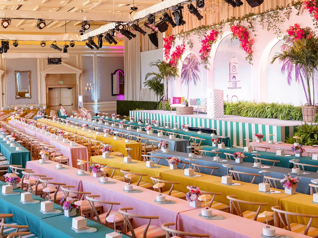 Long tables in rainbow-striped linens set with mini florals and favor boxes beneath a full overhead lighting truss, facing a tropical mural stage with a painted clocktower and bougainvillea.